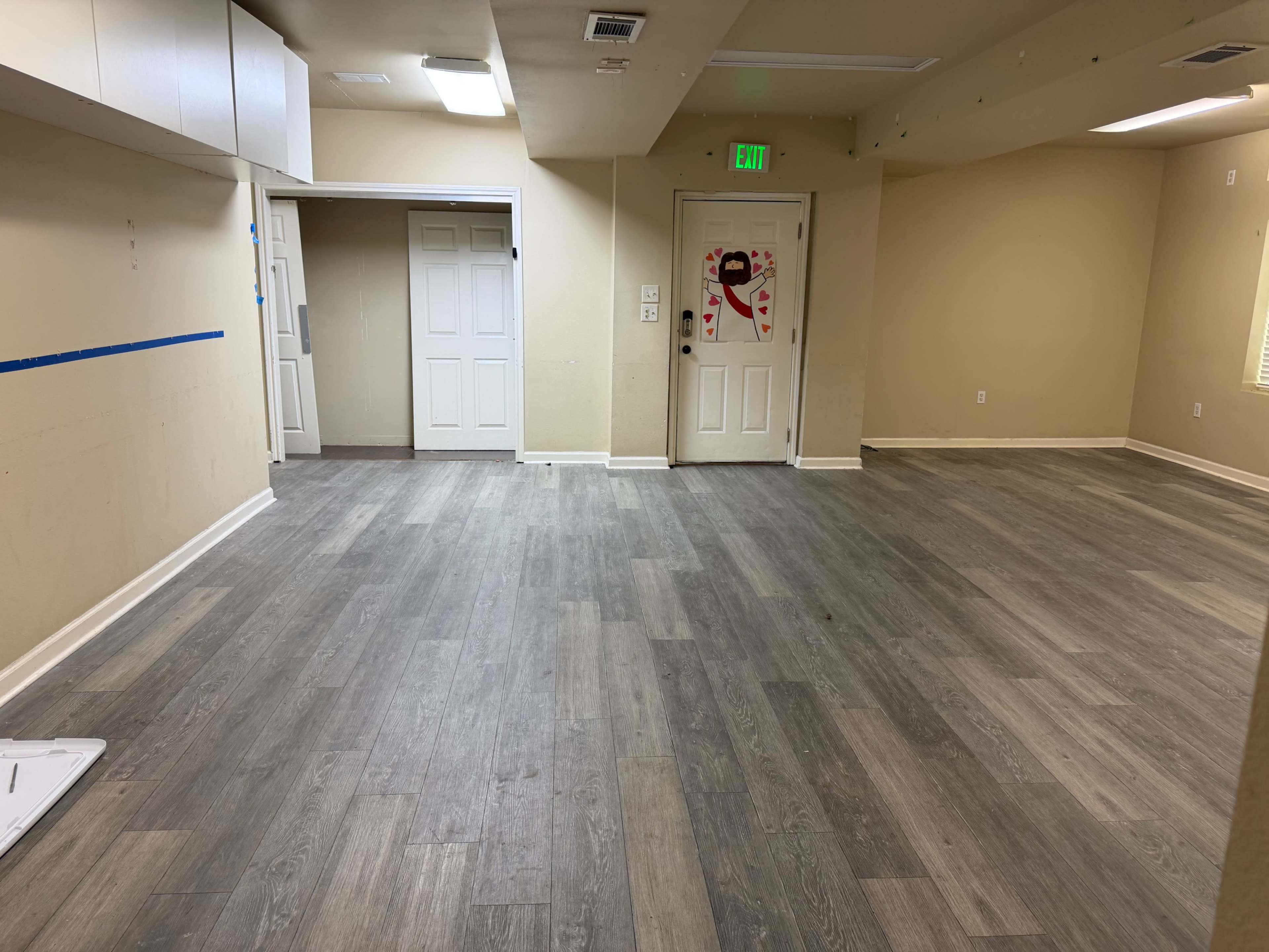 The image shows a spacious, empty room with light-colored walls and gray wooden flooring, featuring closed double doors and a visible strip of blue tape on the wall.