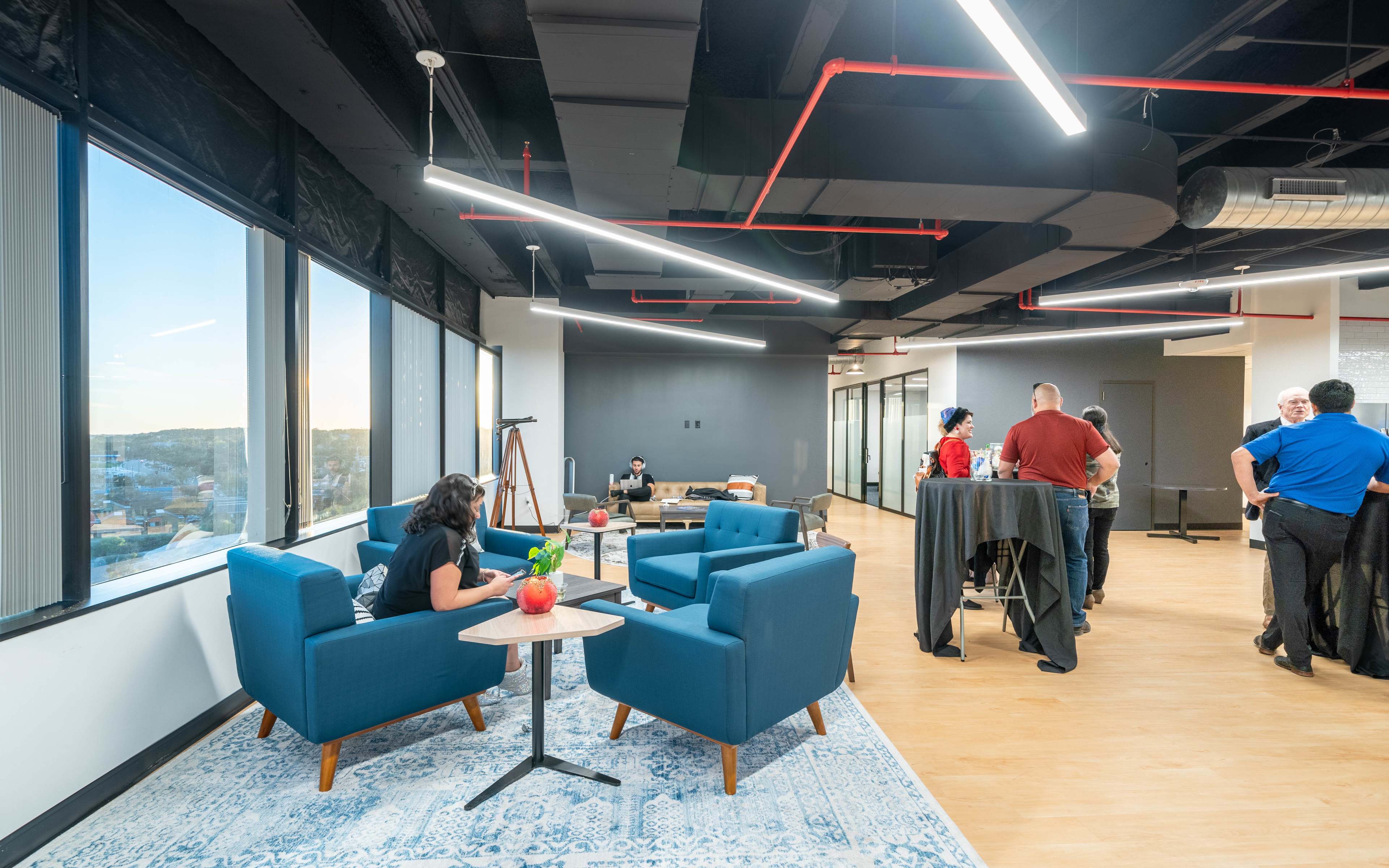 An office space features modern seating areas with blue chairs and a table, while people engage in conversation and work at standing tables.