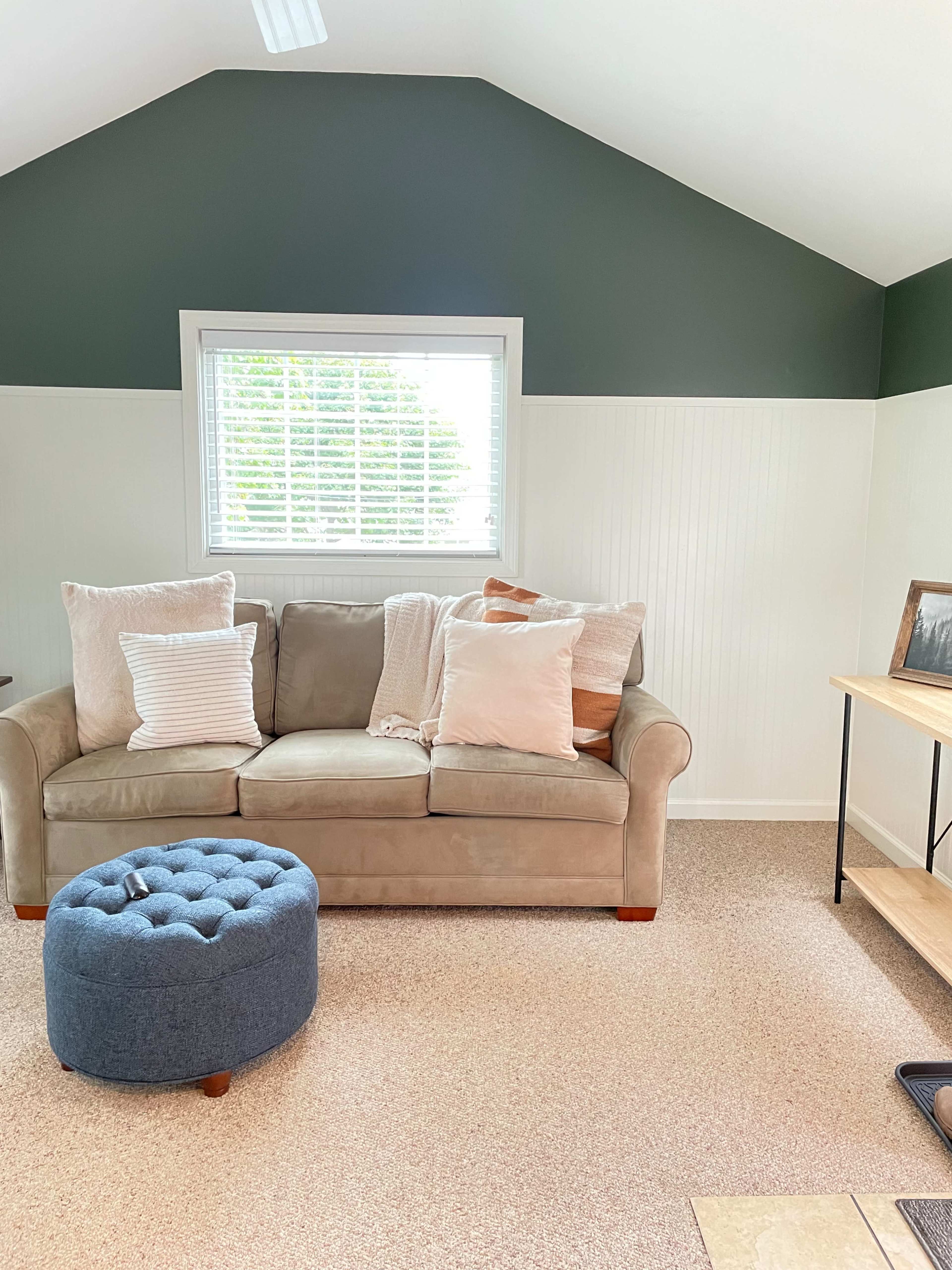 A light-filled room features a beige couch with decorative pillows, a blue ottoman, and a large window framed by white walls and a dark green accent wall.