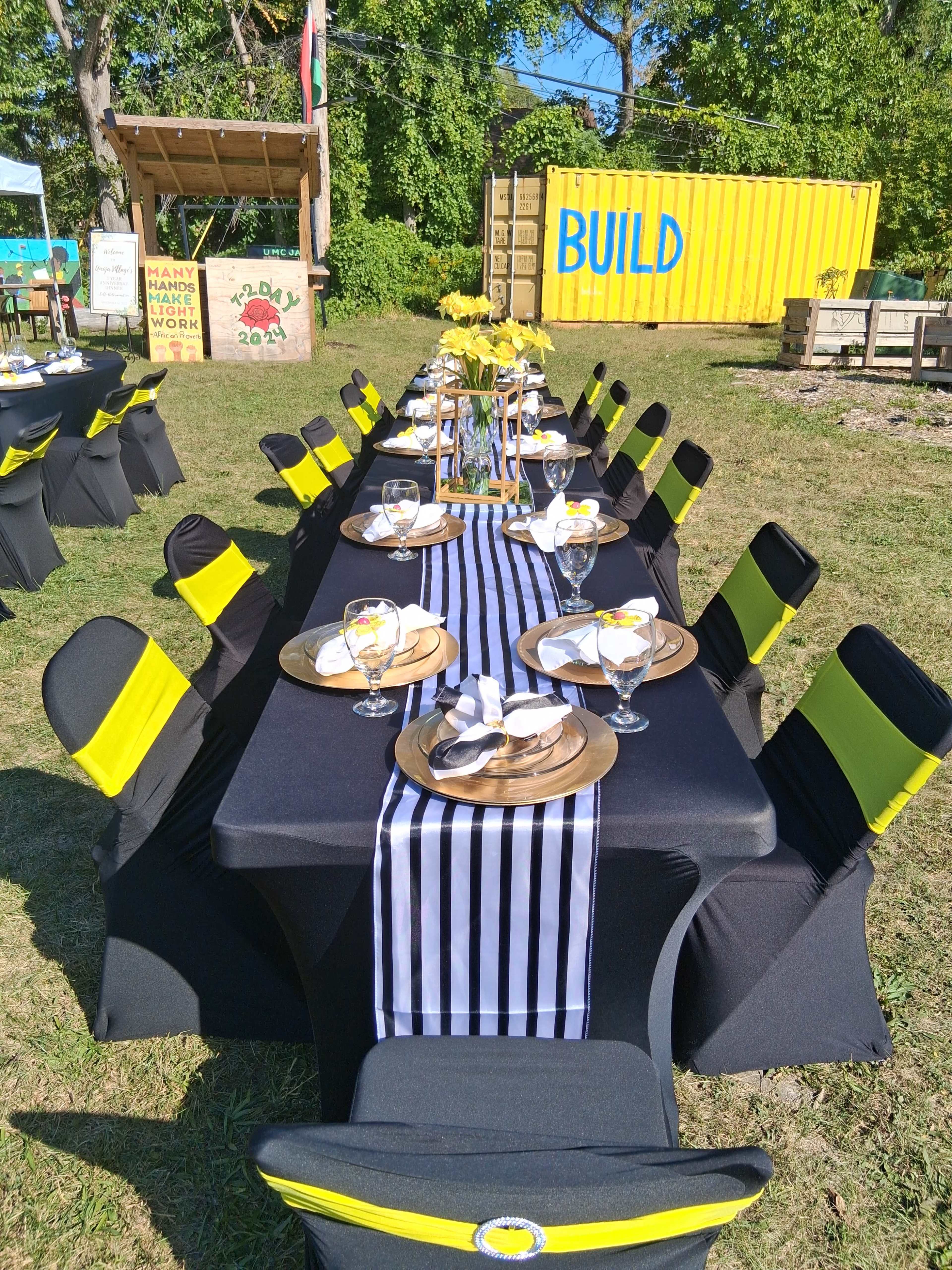 A long, outdoor dining table is set with black and yellow chair covers, striped runners, and decorative plates, surrounded by greenery and shipping containers.