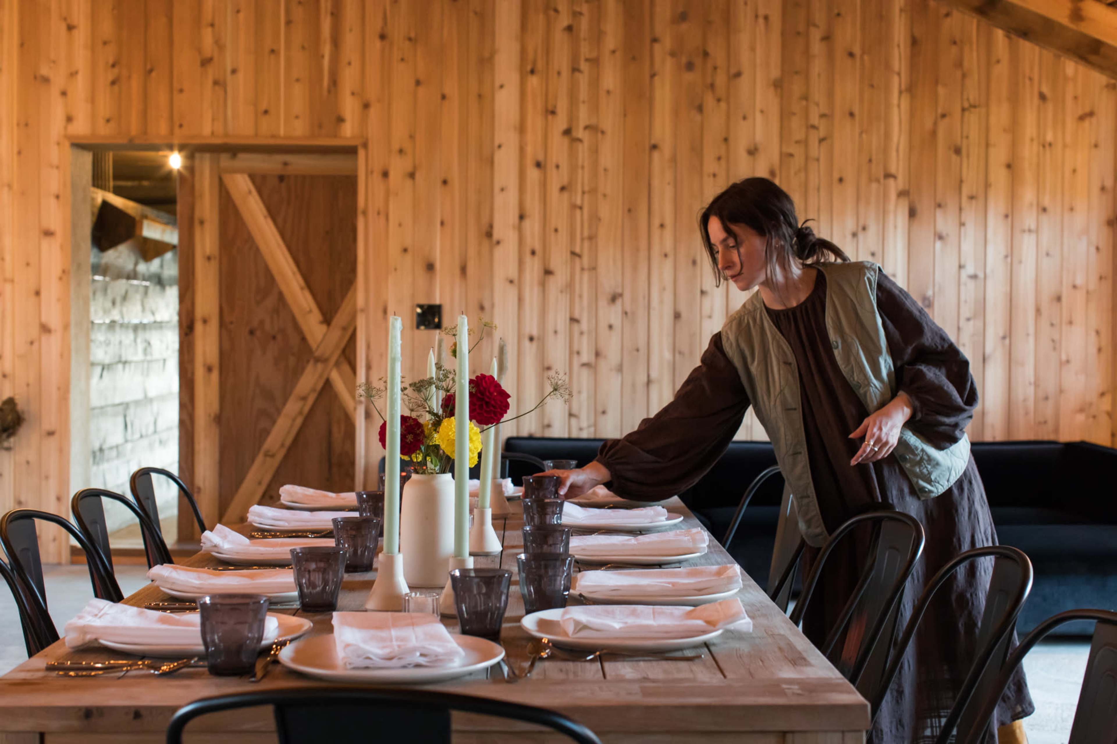 A woman adjusts the table setting in a wooden-paneled dining area with a large table and black chairs.
