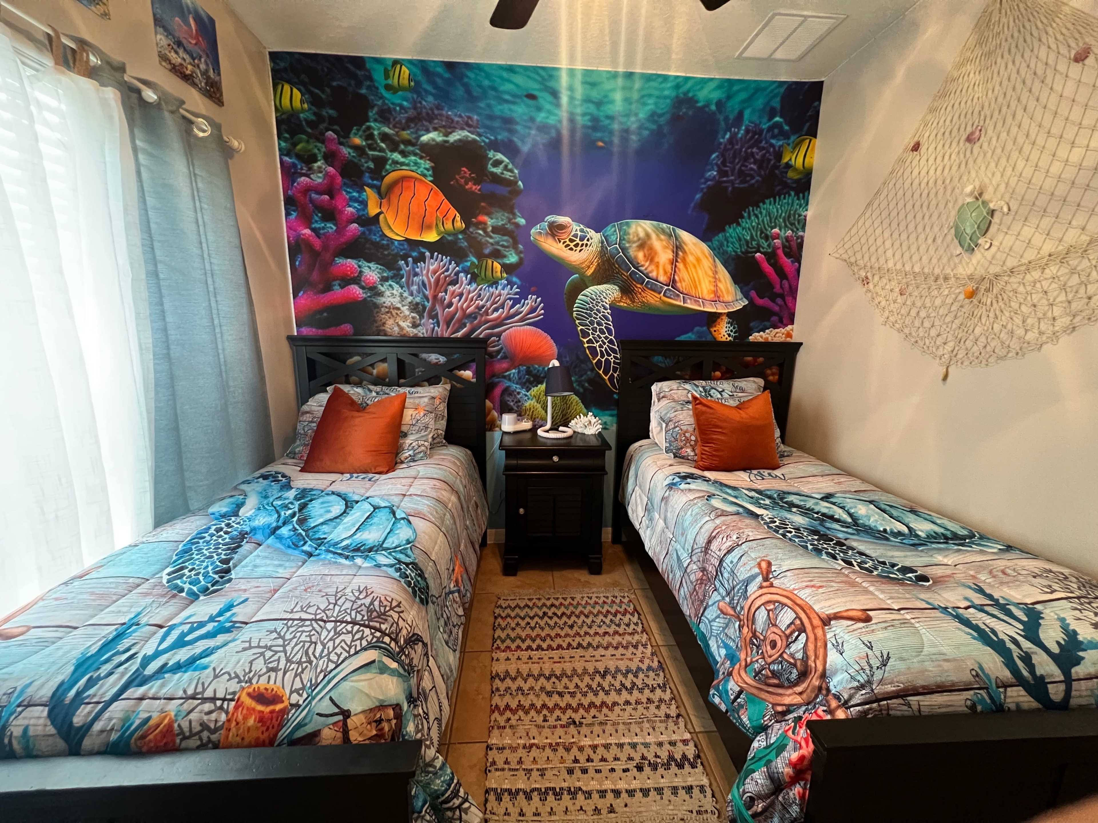 The image shows a colorful underwater-themed bedroom with two twin beds adorned with patterned bedspreads, a sea turtle mural on the wall, and decorative pillows.