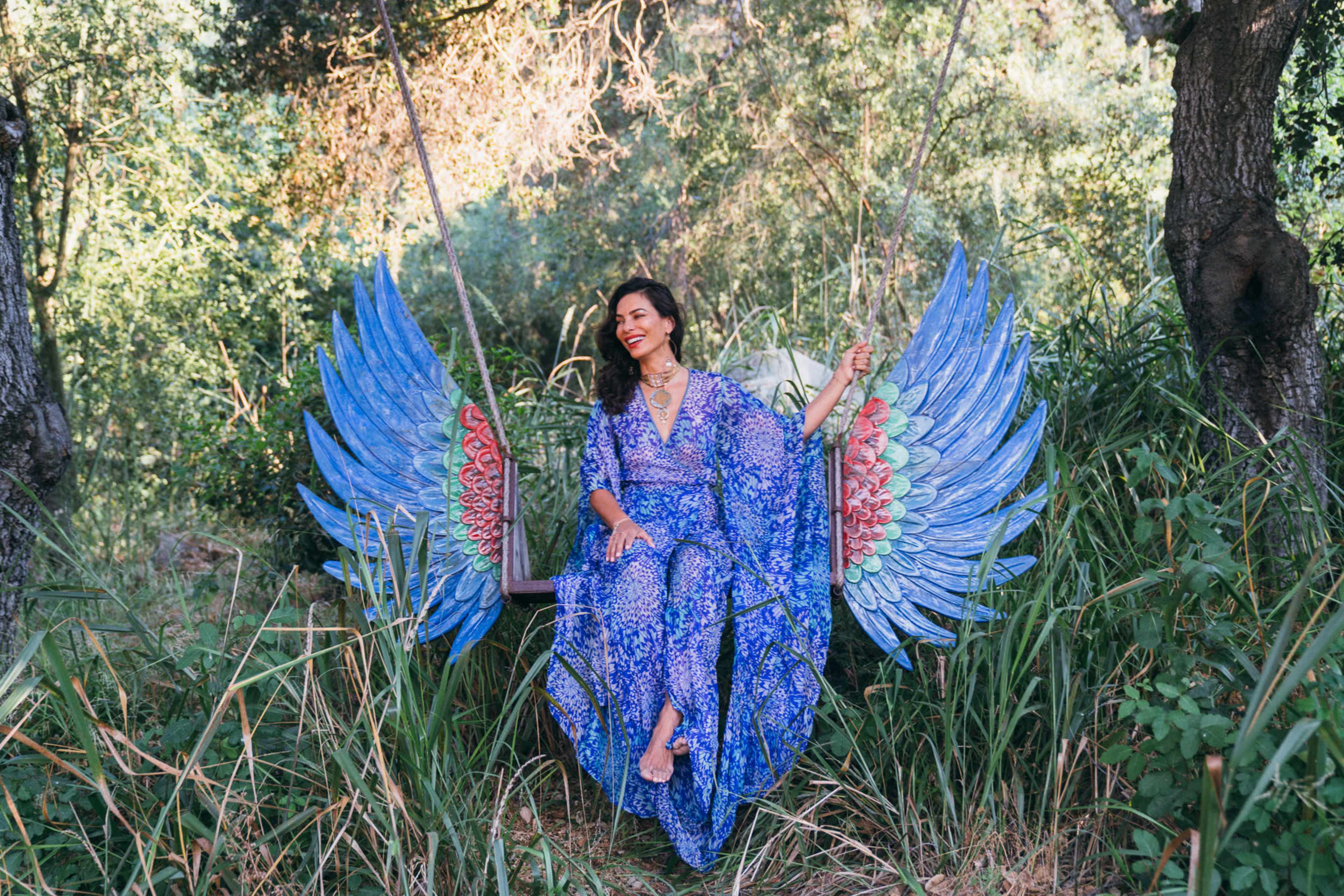 A woman sits on a swing adorned with large, colorful wings in a lush, green forest setting.