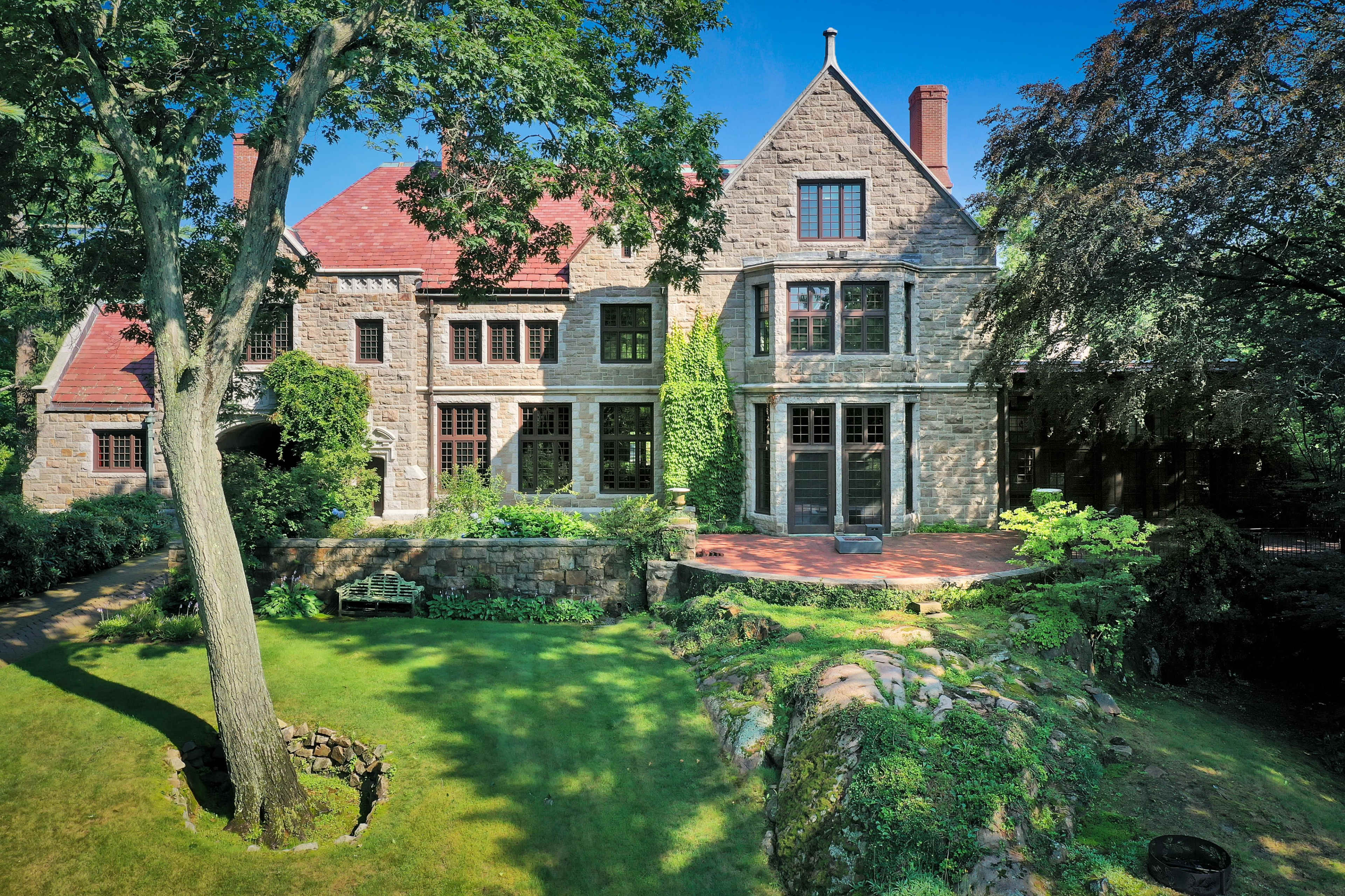 The image shows a large stone house with a red roof, surrounded by lush greenery and a manicured lawn.