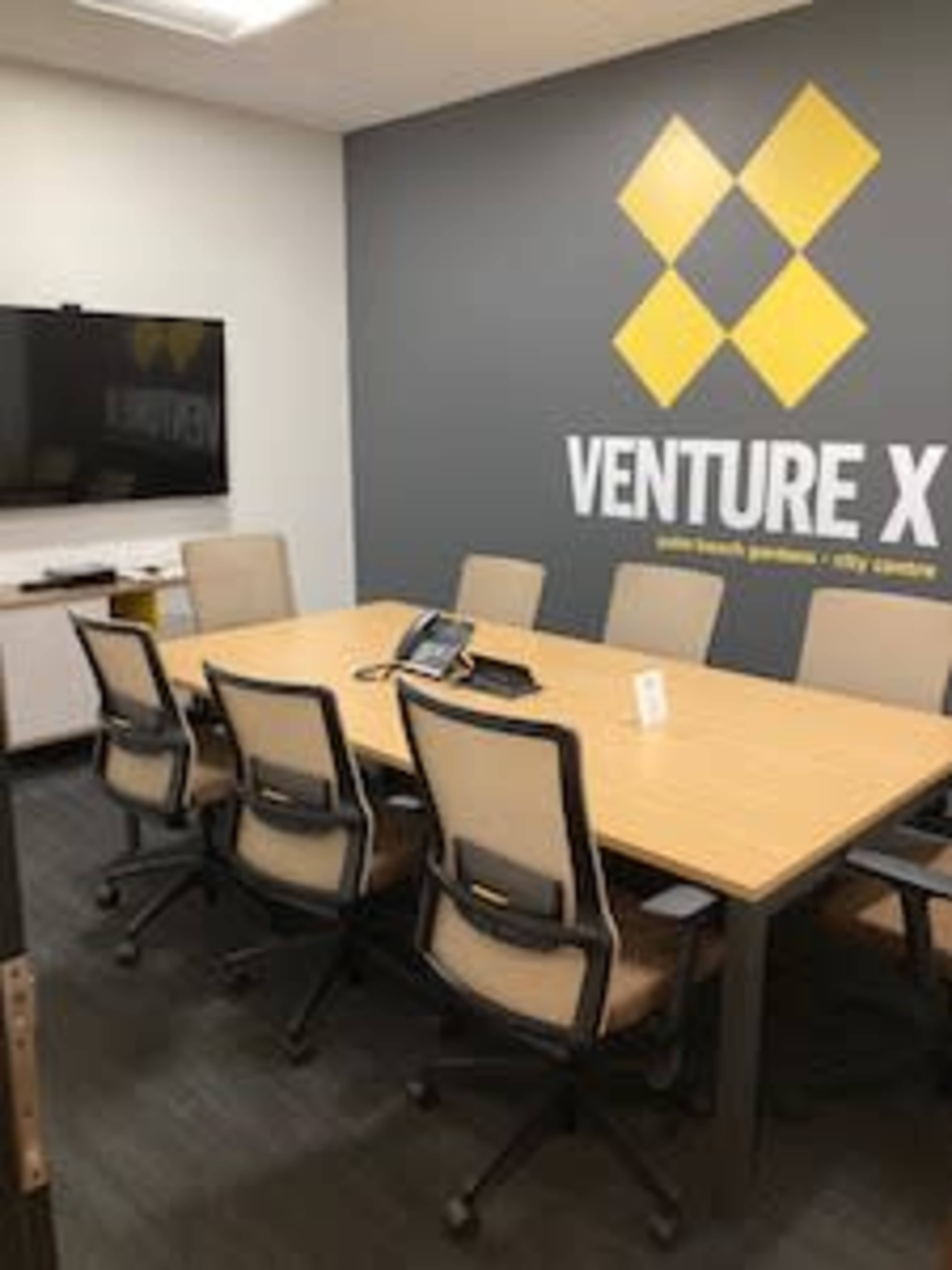 The image shows a conference room featuring a large wooden table surrounded by six chairs, a television mounted on the wall, and a branded wall sign that reads "VENTURE X."