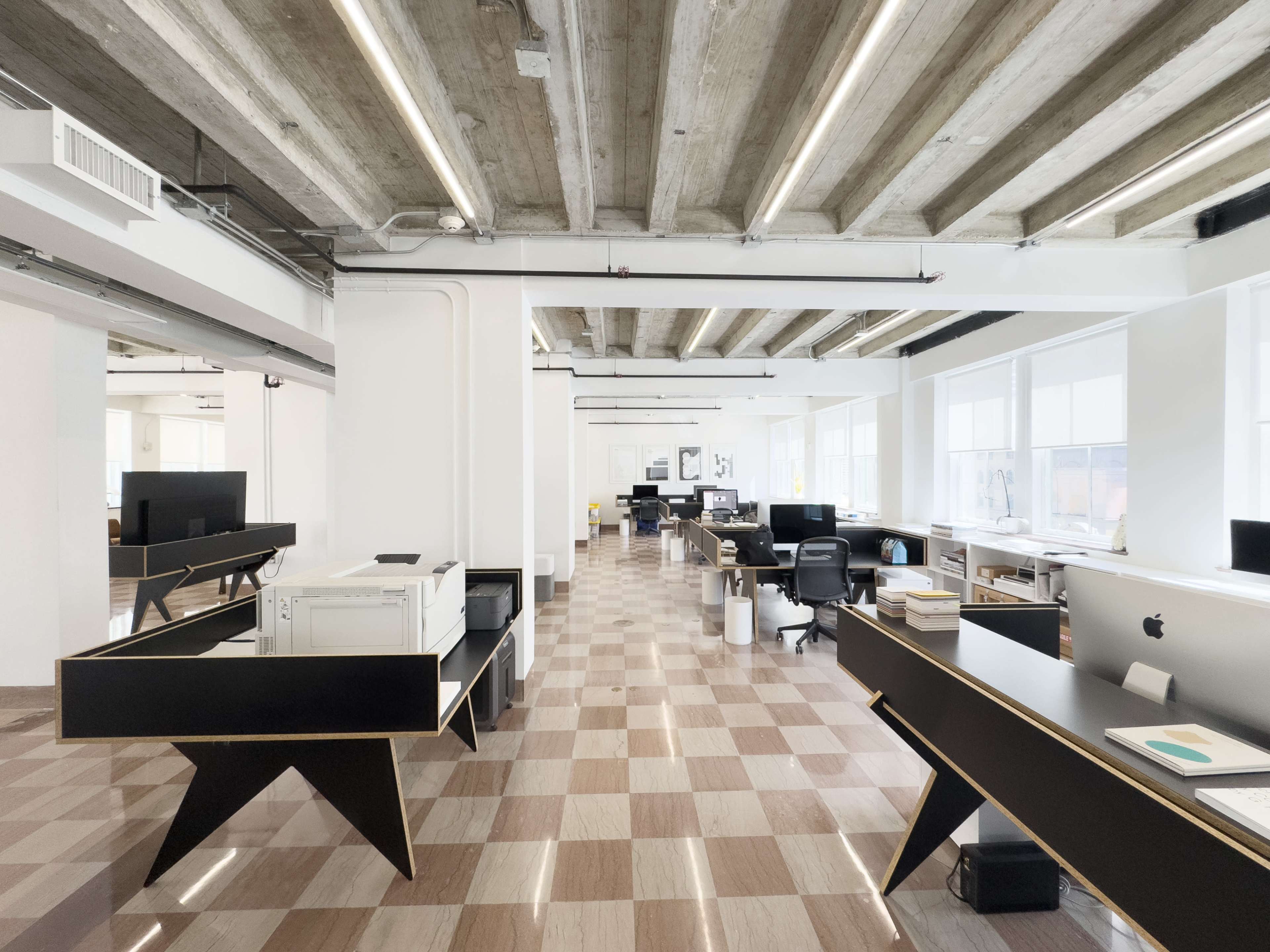 The image shows a modern open office space with large windows, light wooden beams, and desks arranged in a grid layout.