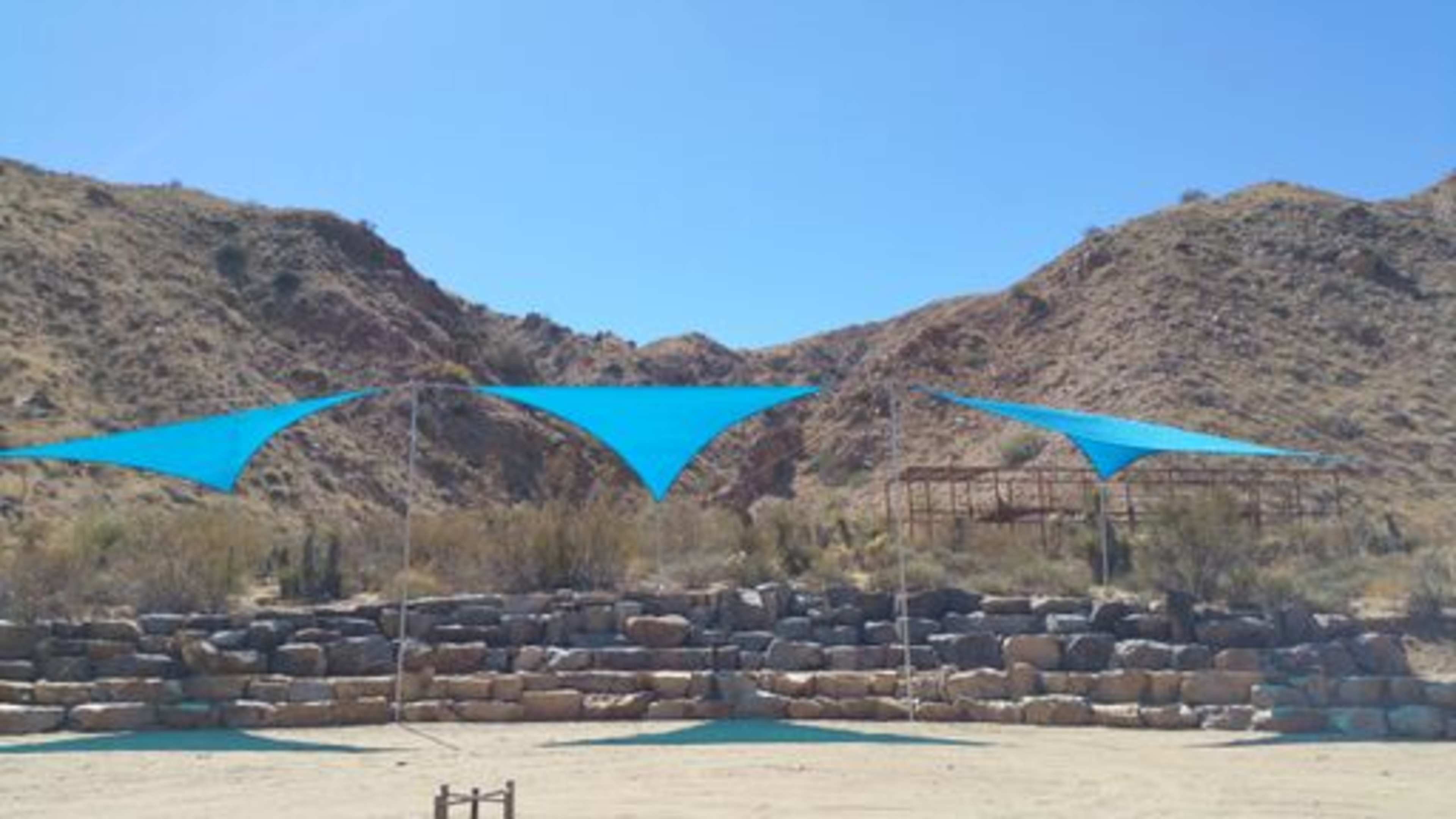 Three large blue shade sails set up in a desert landscape with rocky hills in the background.