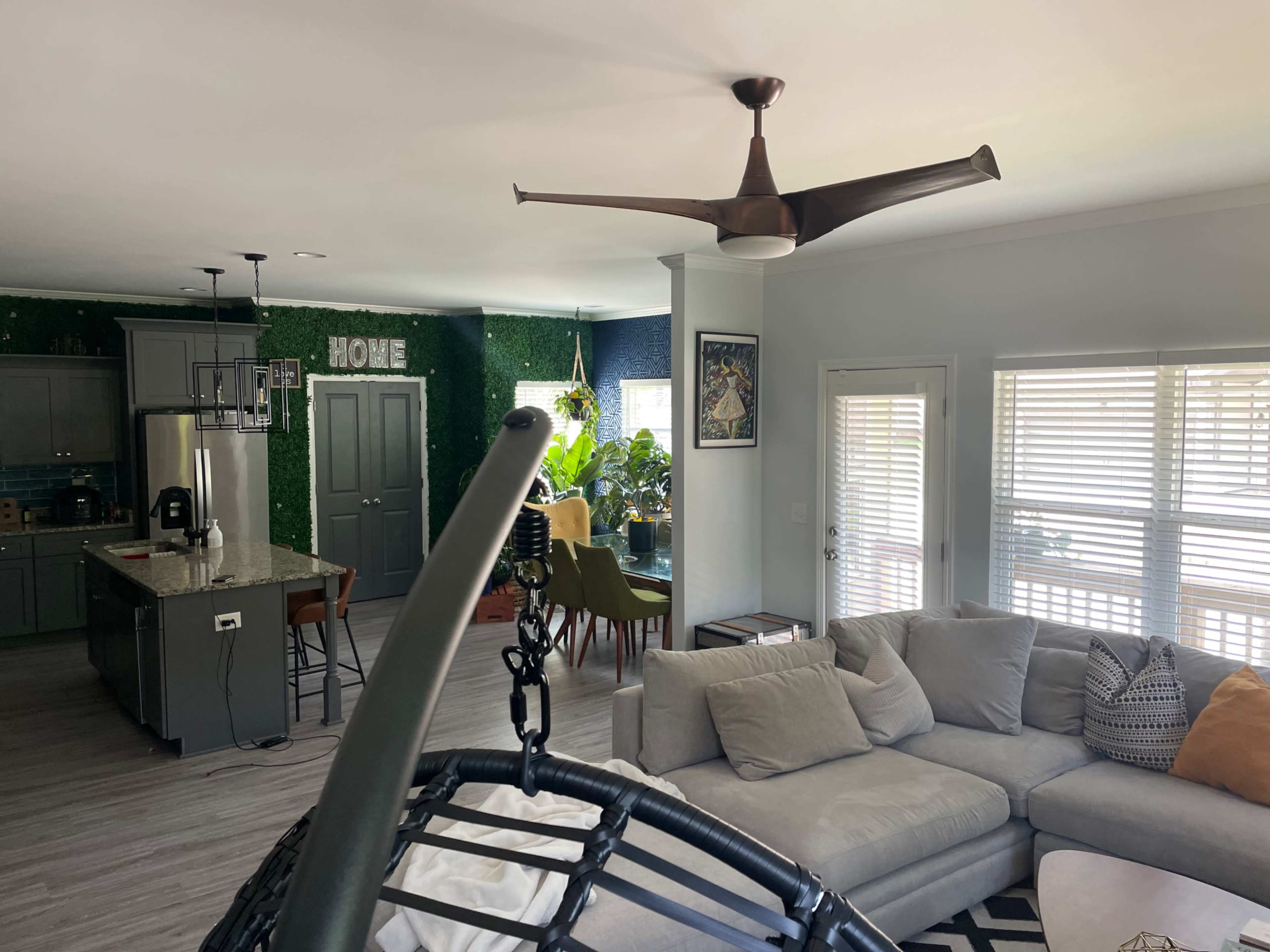 The image shows a modern living room with a large sectional sofa, a hanging chair, and a kitchen space featuring green accents and houseplants.