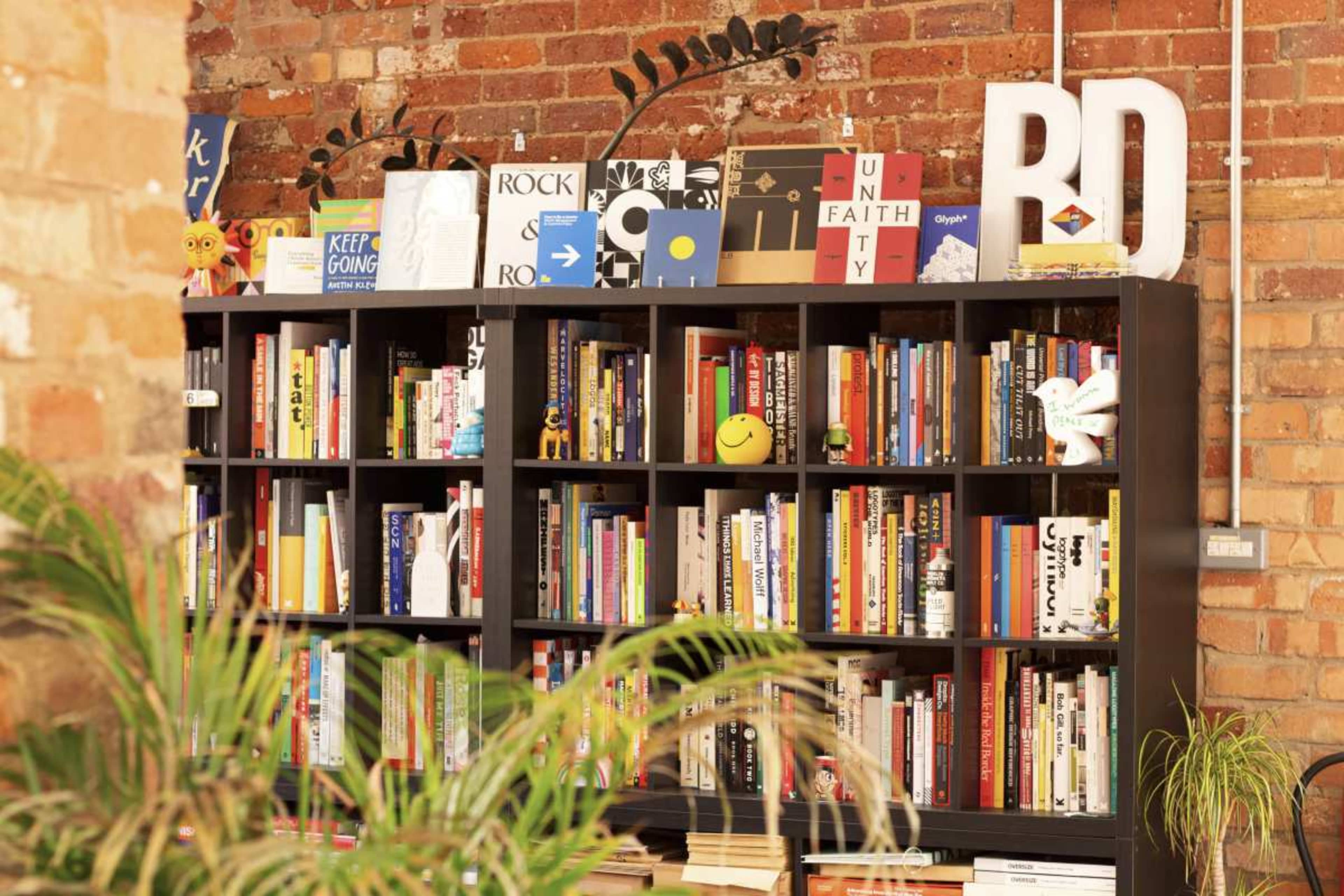 The image shows a black bookshelf filled with various books and decorative objects, positioned against a brick wall.