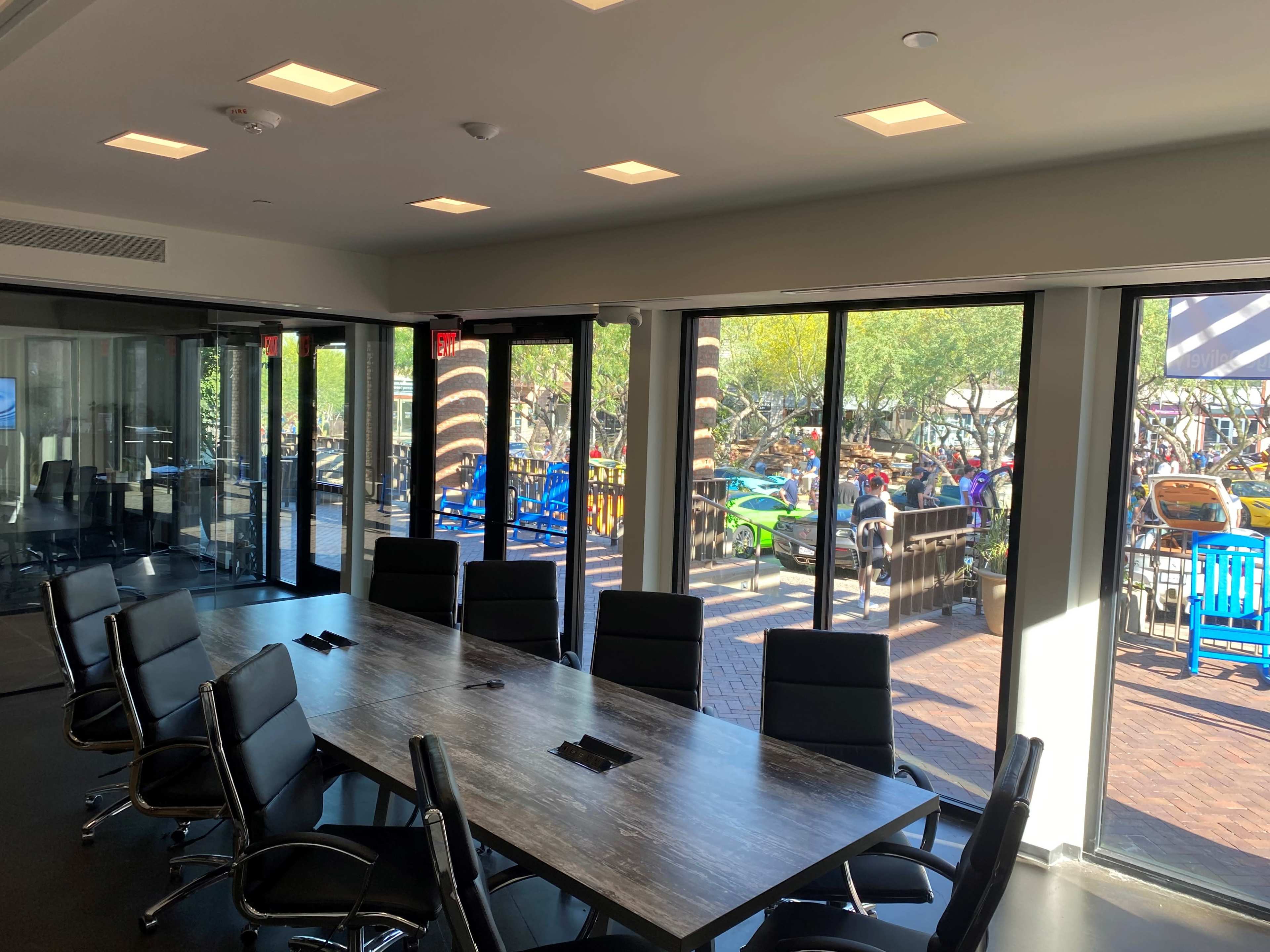 A conference room with a long table and black chairs has large glass windows overlooking an outdoor area with colorful recreational equipment.
