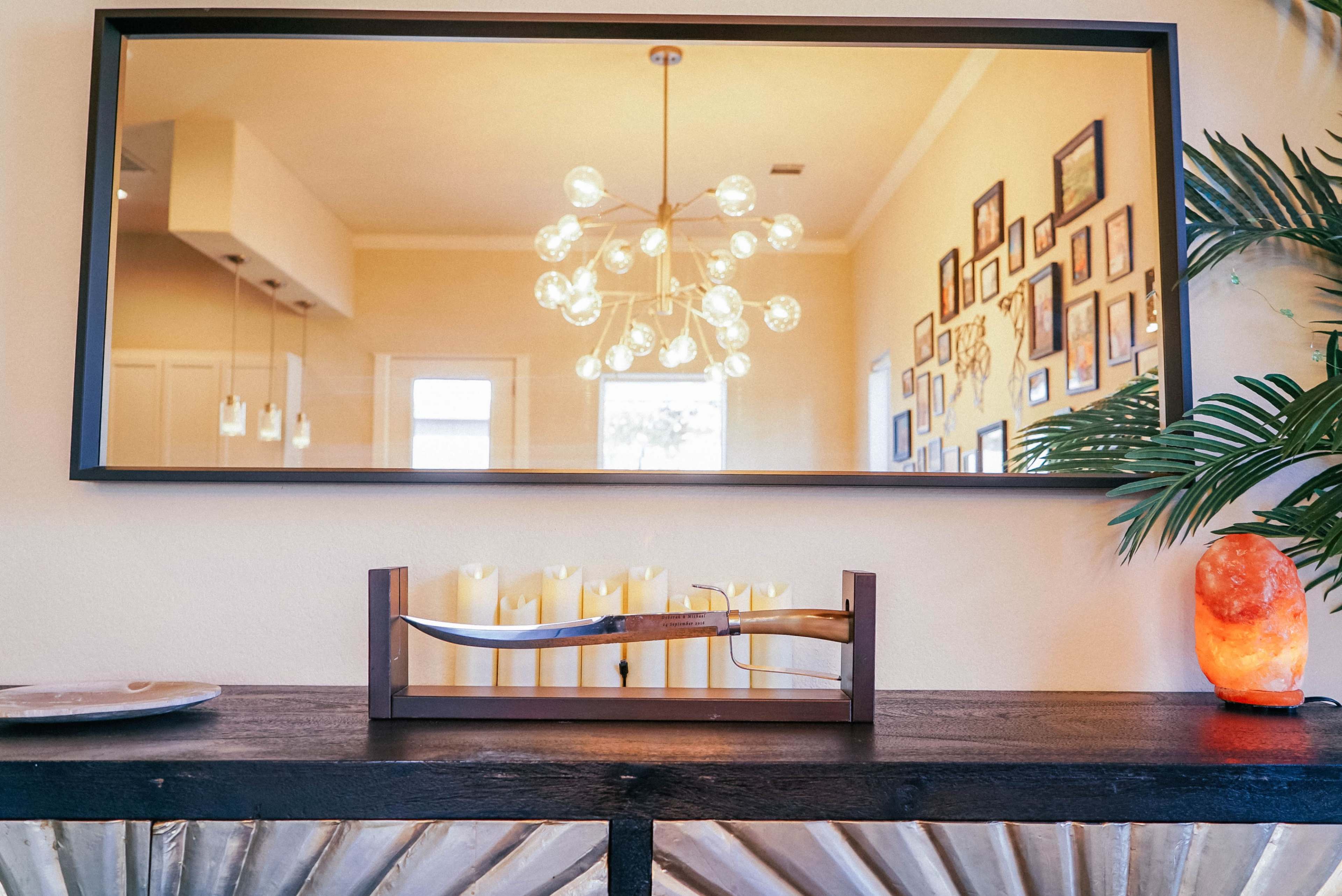 A wooden shelf displays a sword under a mirror reflecting a decorated wall and a light fixture in the background.