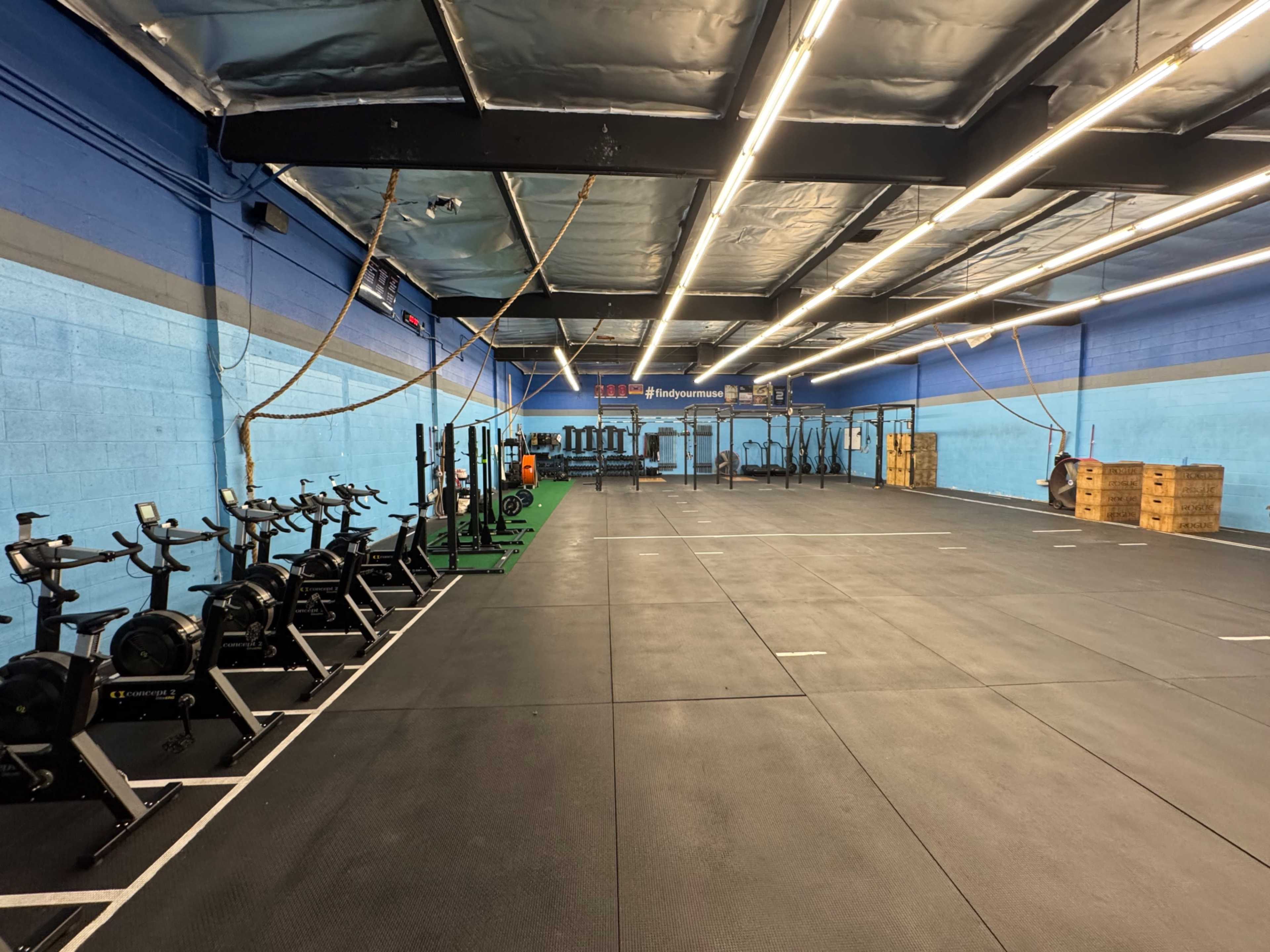 The image shows a spacious gym with rows of exercise equipment, including stationary bikes, located on a rubber floor beneath bright overhead lights.