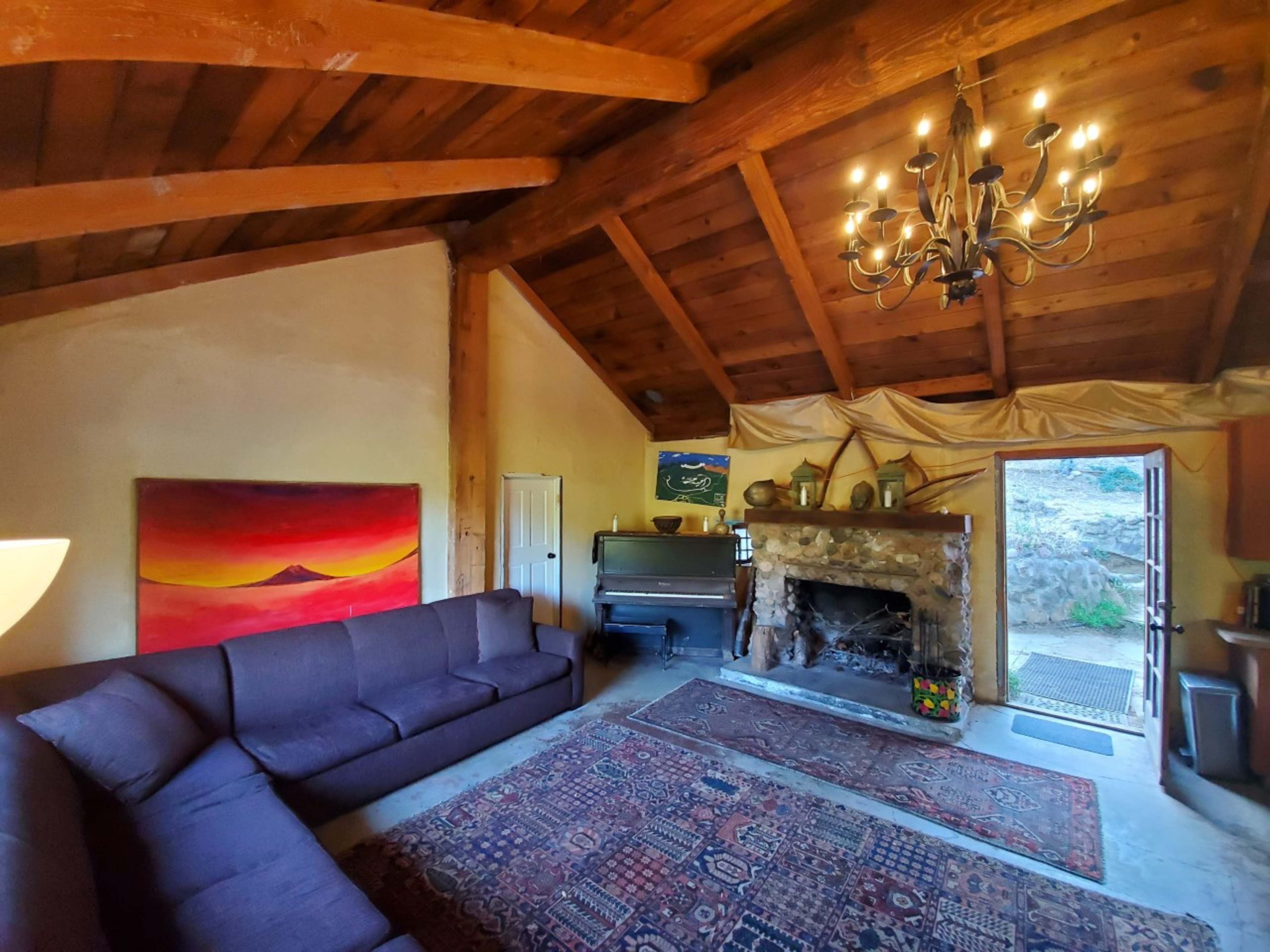 A cozy living room with a stone fireplace, a large dark sofa, and wooden beams on the ceiling, leading to an outdoor area.
