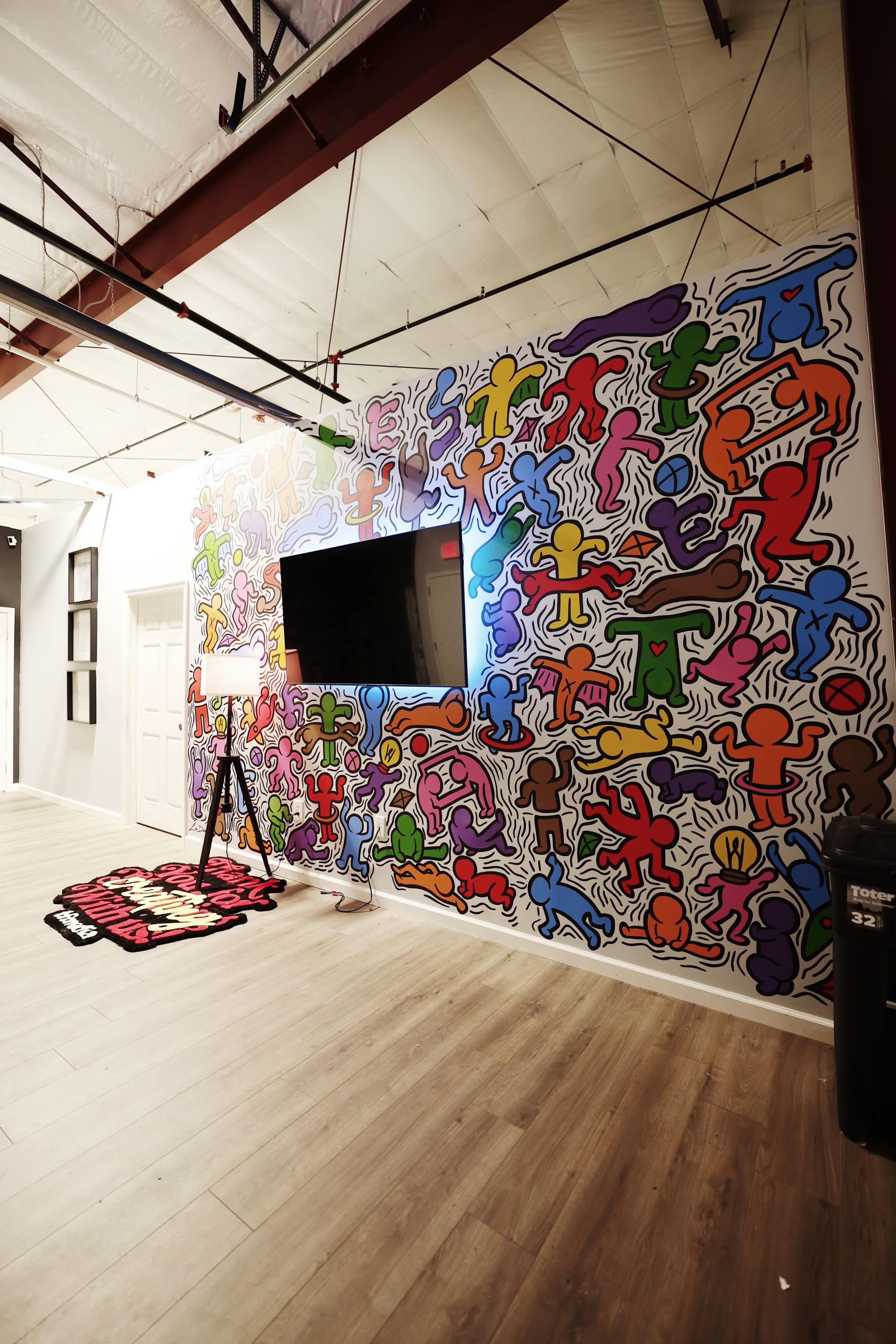 A brightly painted wall covered with colorful abstract figures beside a flat-screen television in a spacious room with wooden flooring.