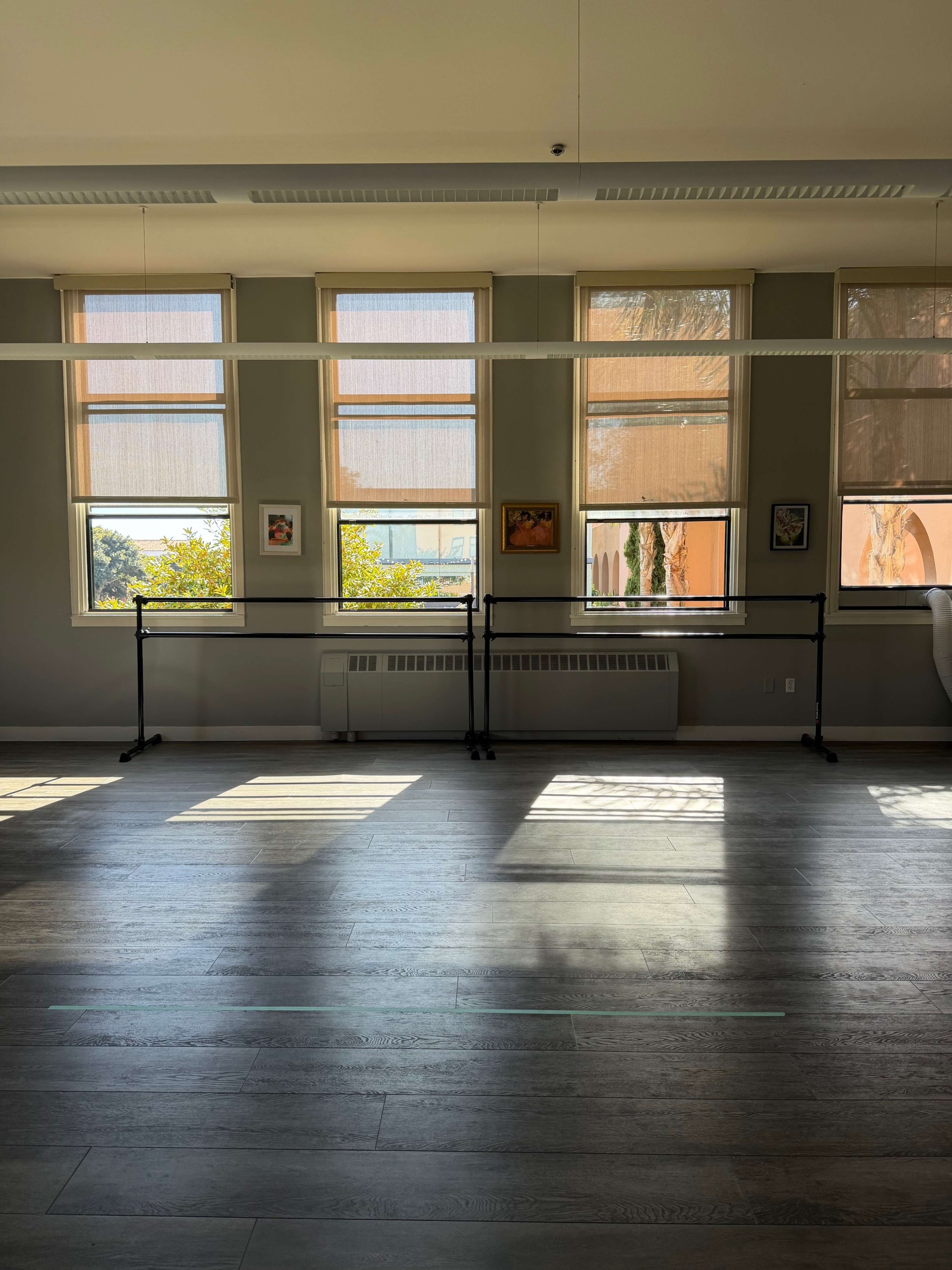 The image shows a bright room with wooden flooring, featuring large windows adorned with shades, and two ballet barres positioned along one wall.