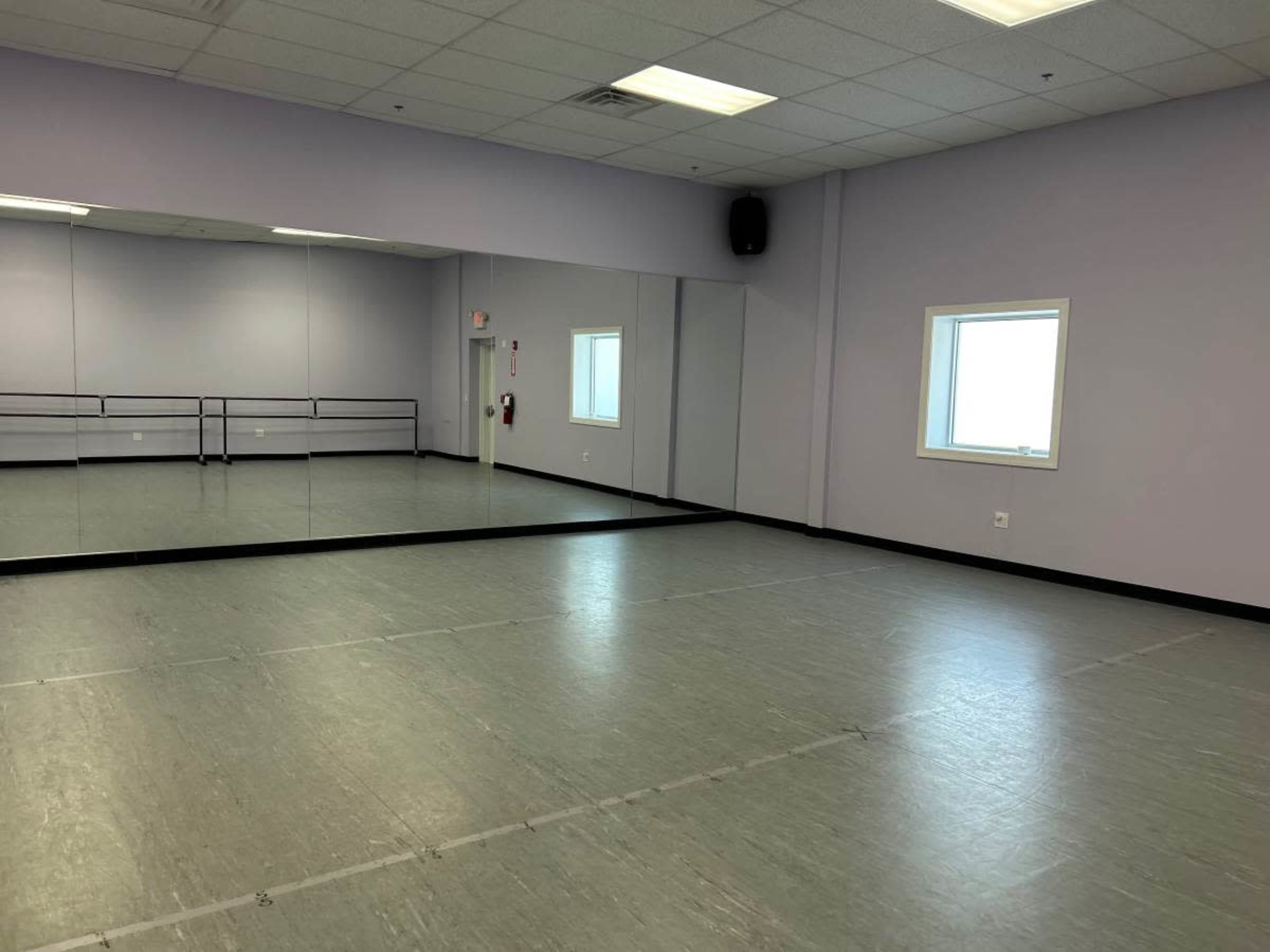 The image shows a spacious dance studio with a mirrored wall and a window, featuring a gray floor and purple walls.