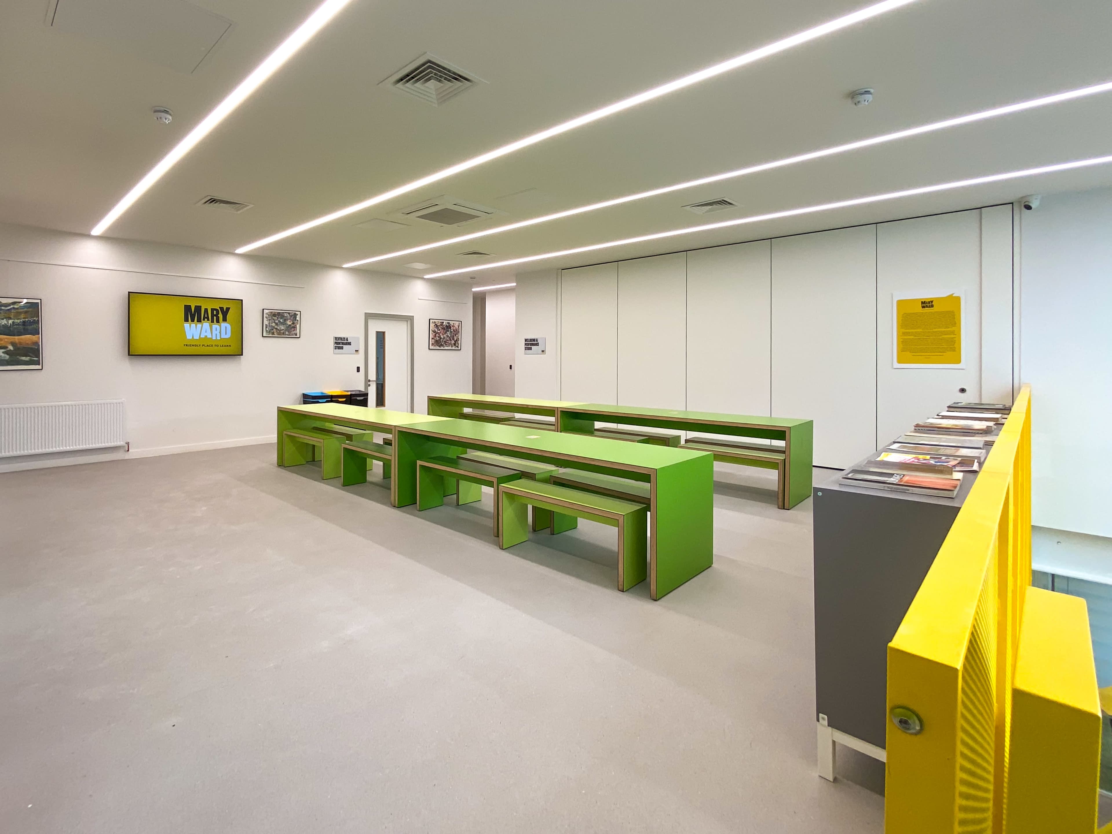 The image shows a modern, minimalist room with green benches arranged in rows, a yellow barrier, and walls displaying various artworks.