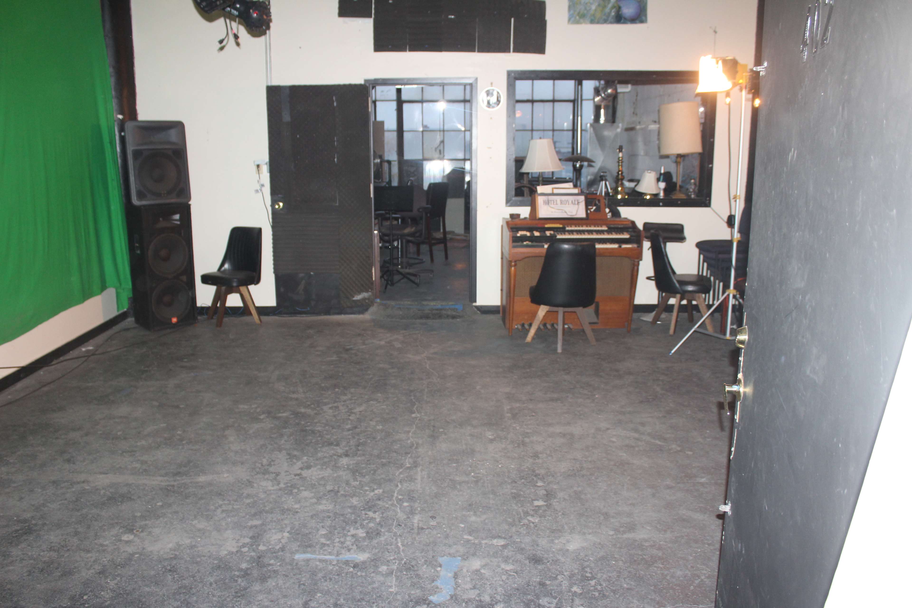 The image shows an empty room with a green screen, a keyboard on a wooden table, and various chairs positioned against the walls.