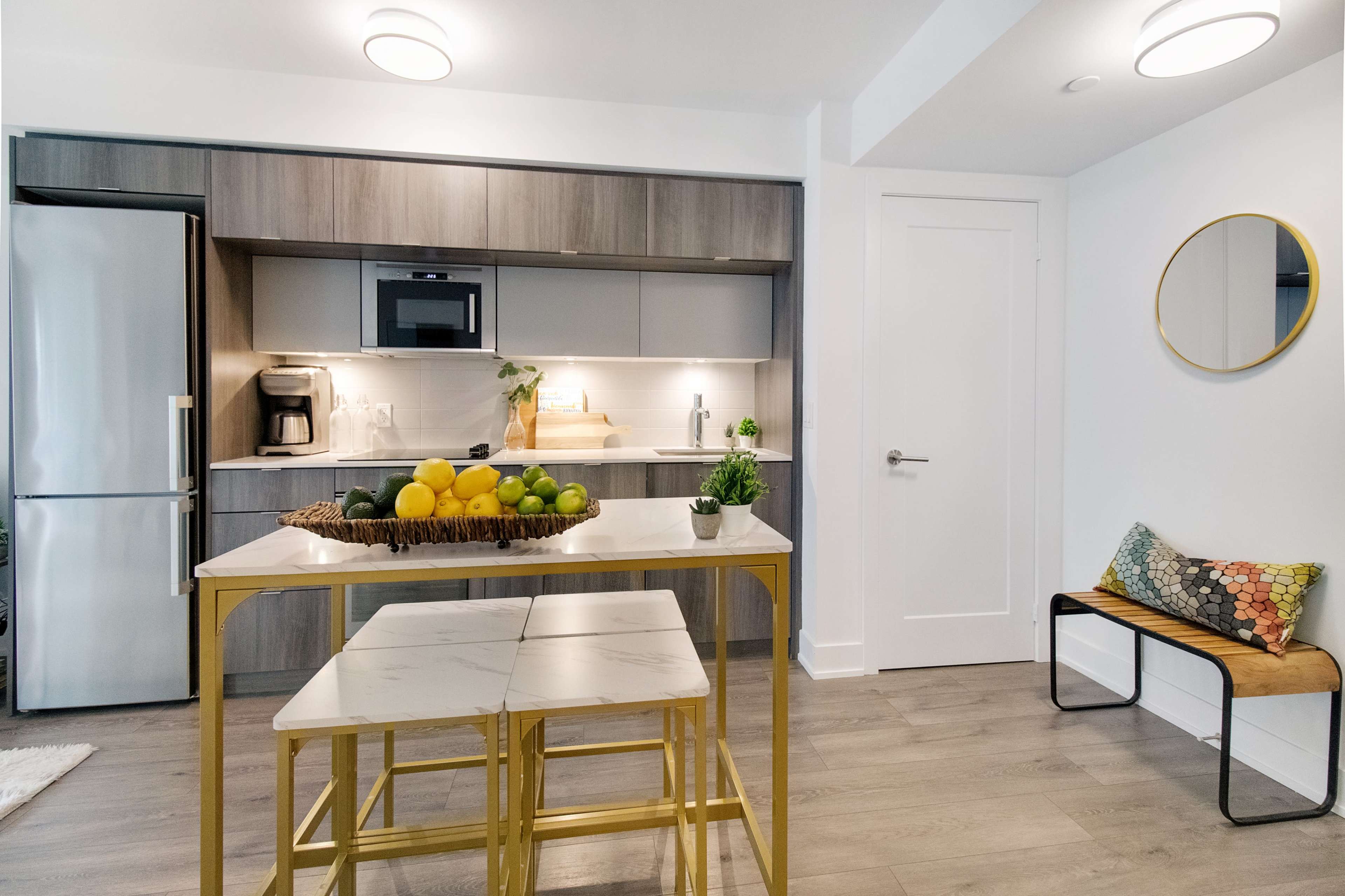 A modern kitchen features a stainless steel refrigerator, gray cabinetry, and a decorative fruit bowl on a gold table with two stools.