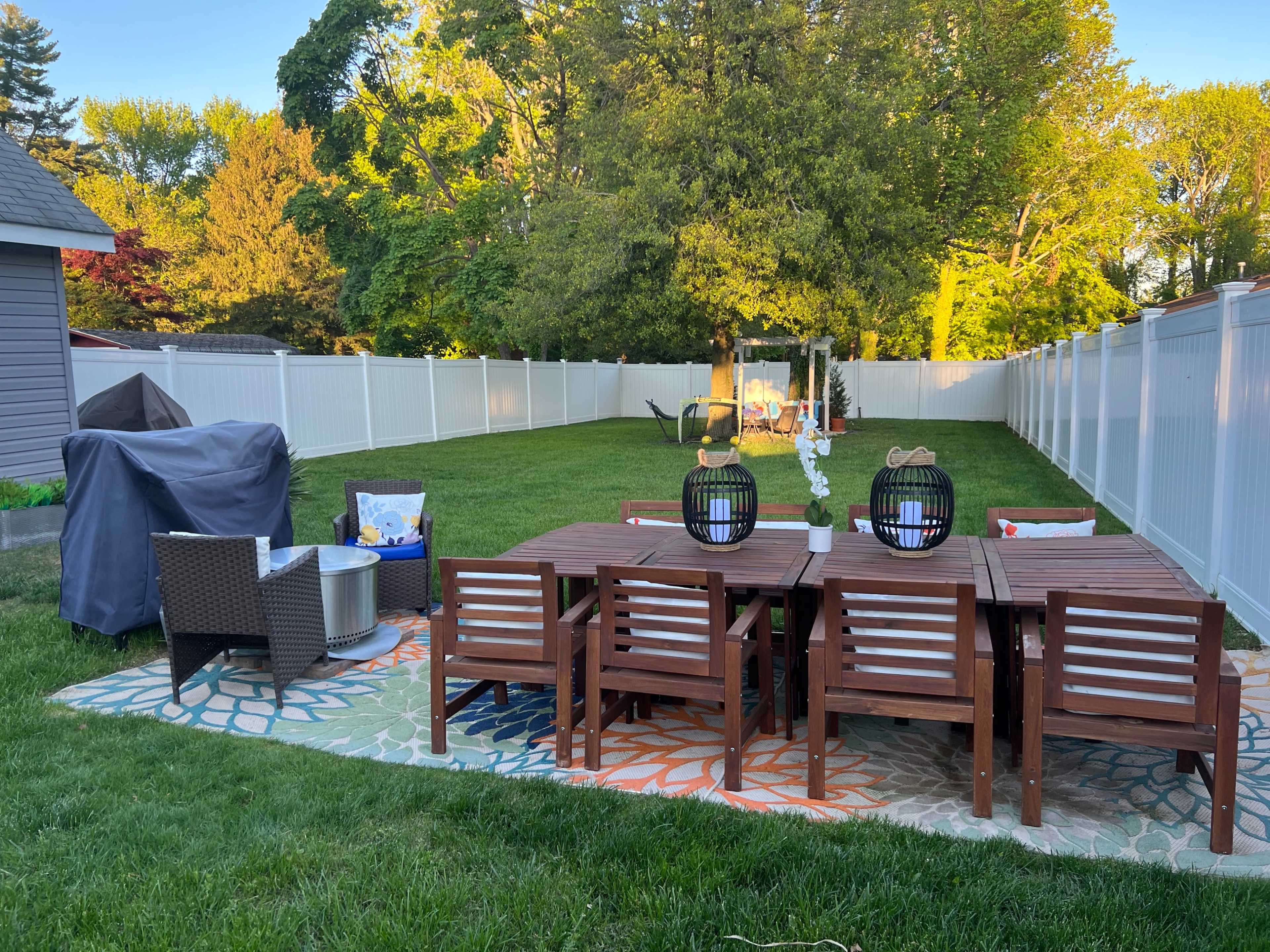 An outdoor patio area with a wooden dining table and chairs, a rug, a grill covered with a tarp, and a lush green lawn enclosed by a white fence.