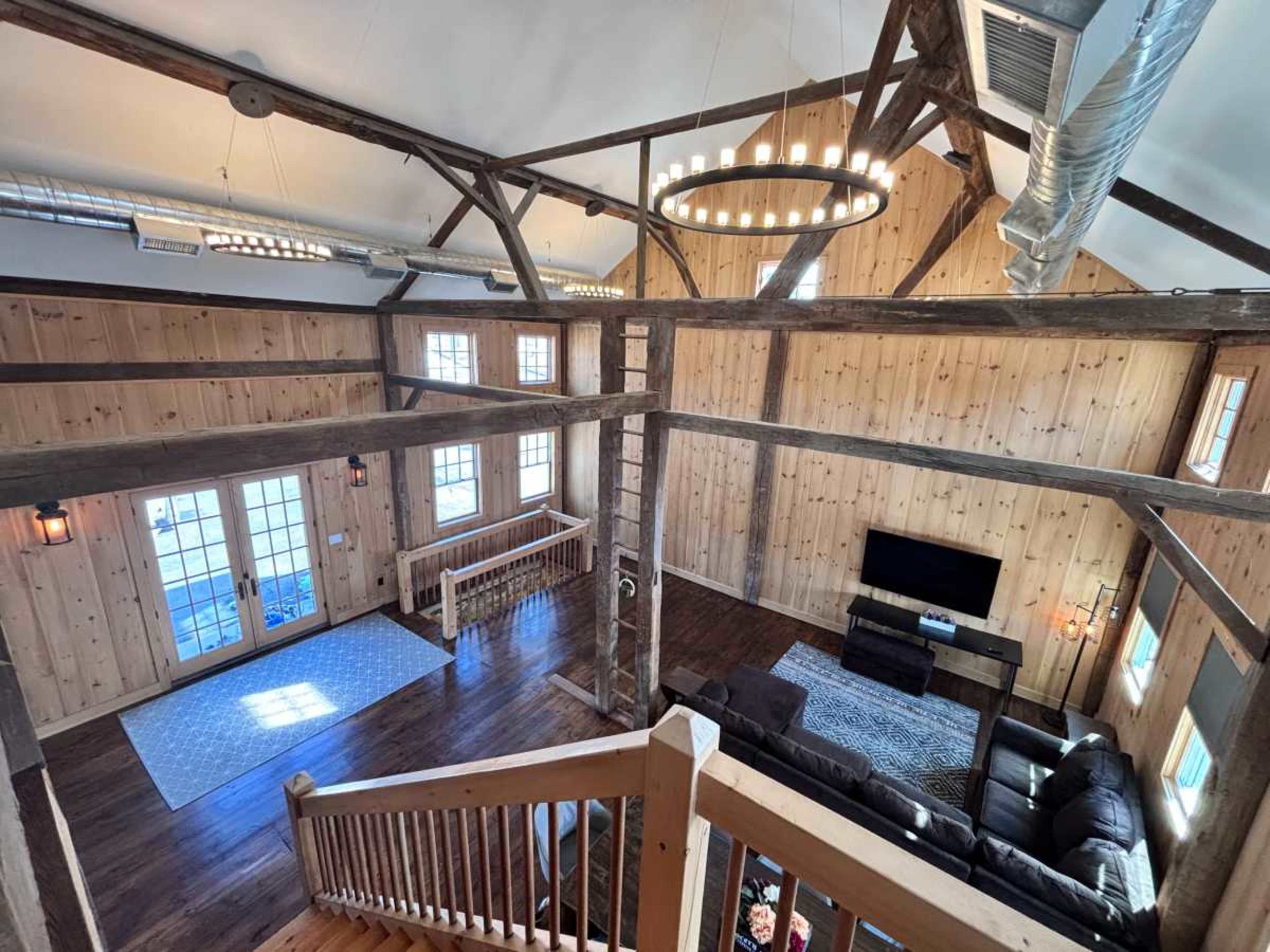 The image shows the interior of a spacious wooden cabin with exposed beams, a large living area, and a staircase leading down to the lower level.