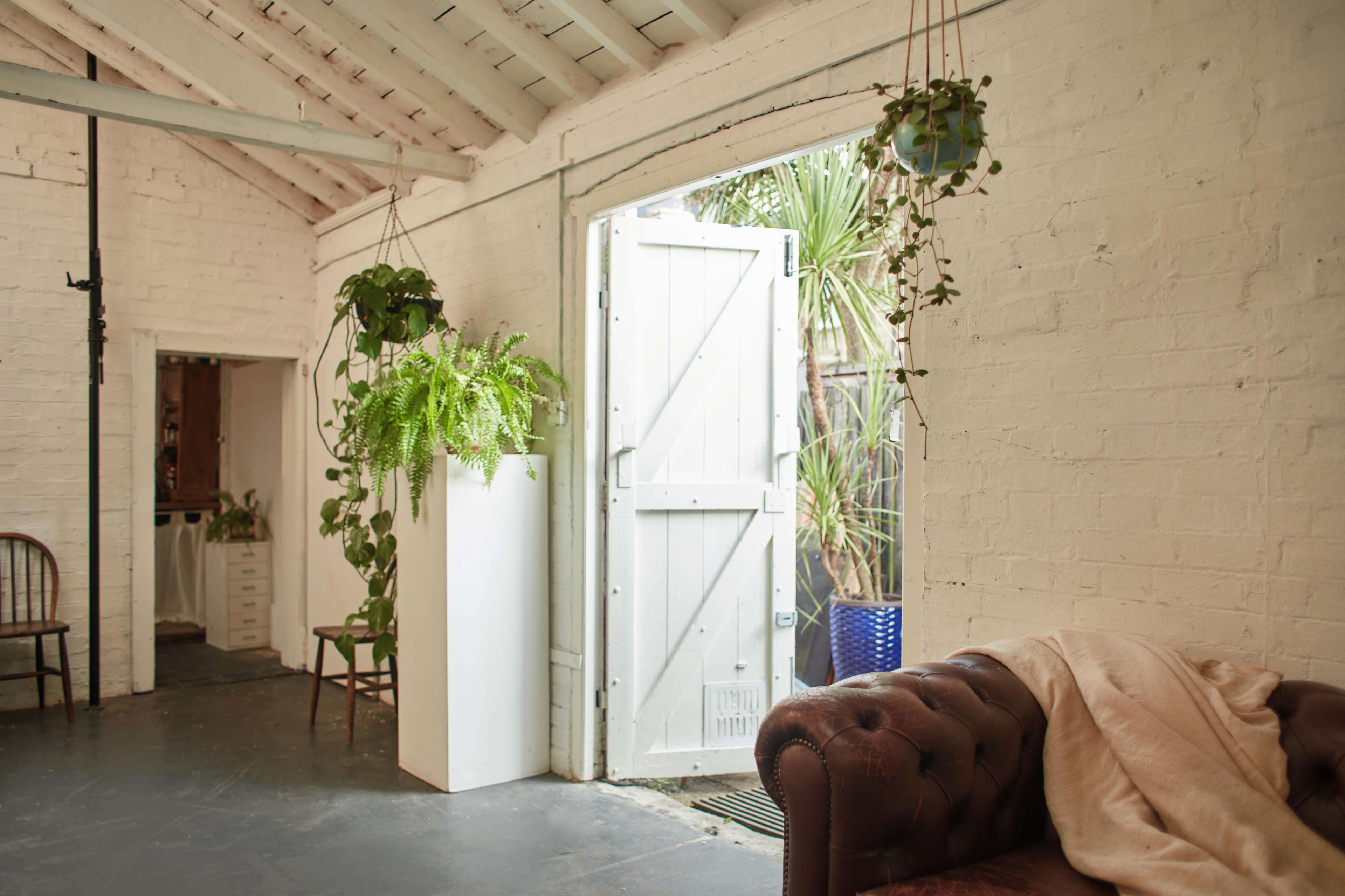 The image shows a bright, airy interior space featuring a white brick wall, a brown leather couch, and an open door leading to a garden with hanging plants.