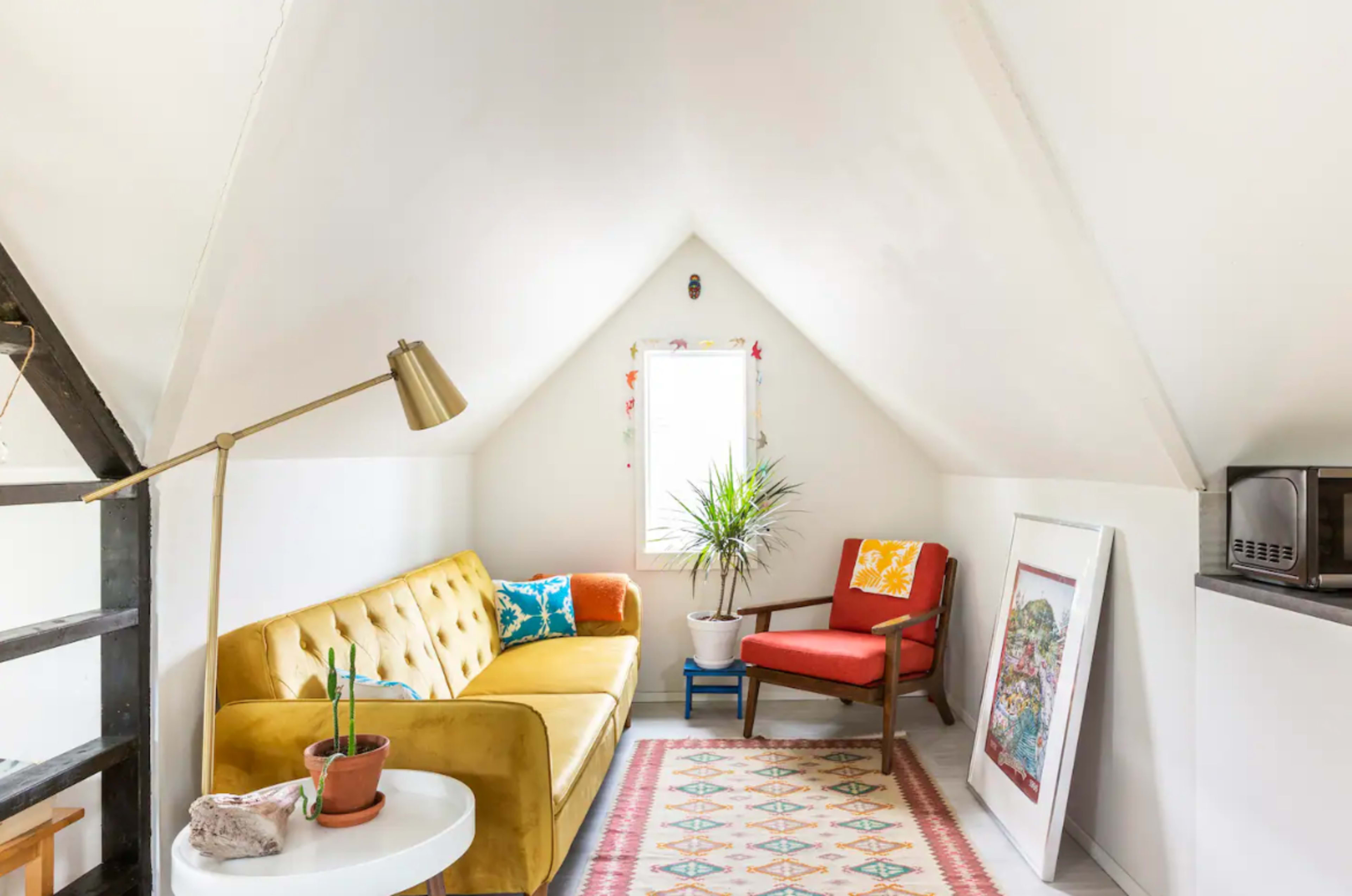 A cozy loft space features a yellow sofa, a red armchair, a potted plant, and artwork, all under a sloped ceiling.