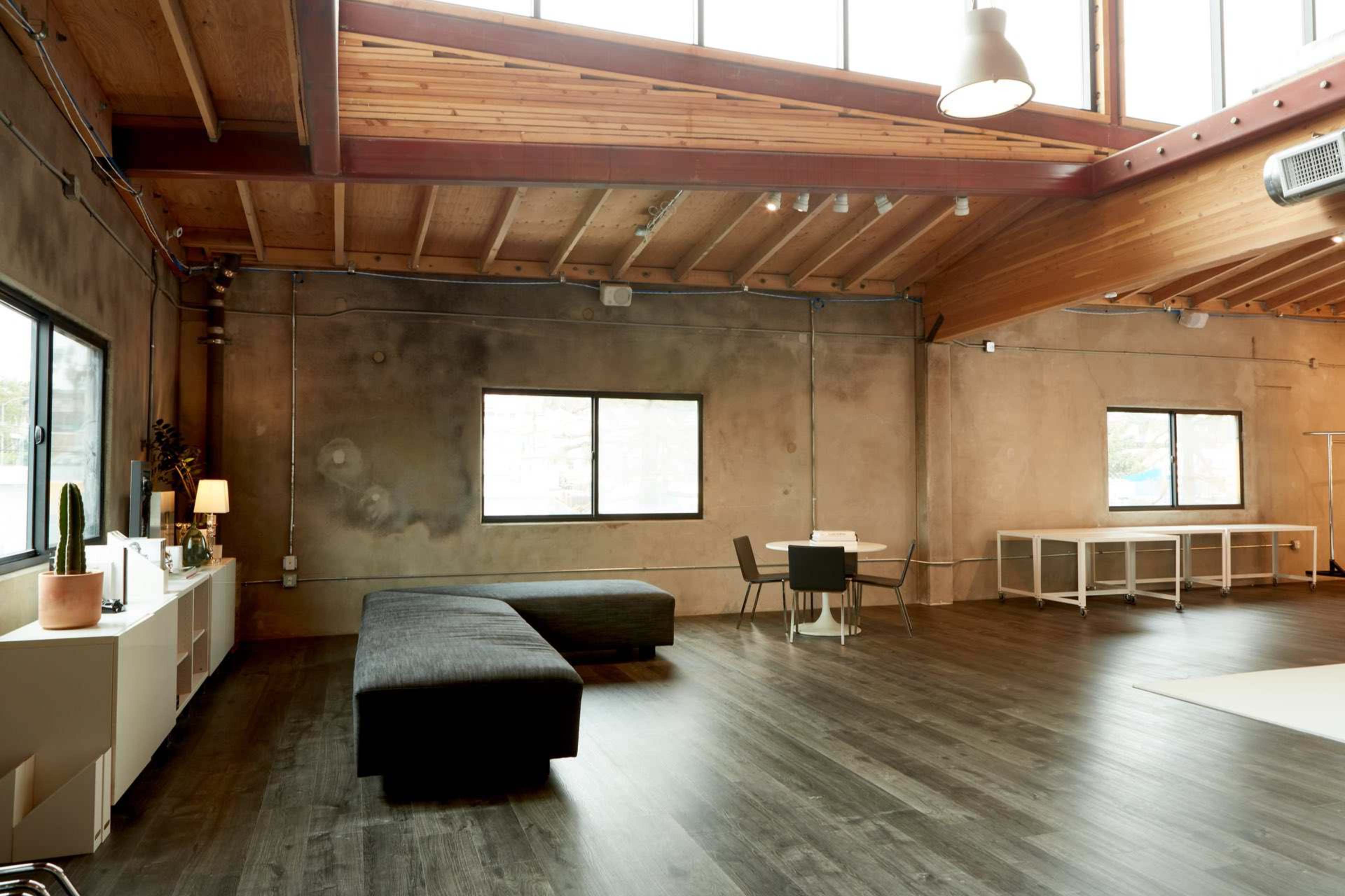 A spacious room with wooden beams, large windows, a dark sofa, a dining table with chairs, and a minimalist aesthetic.