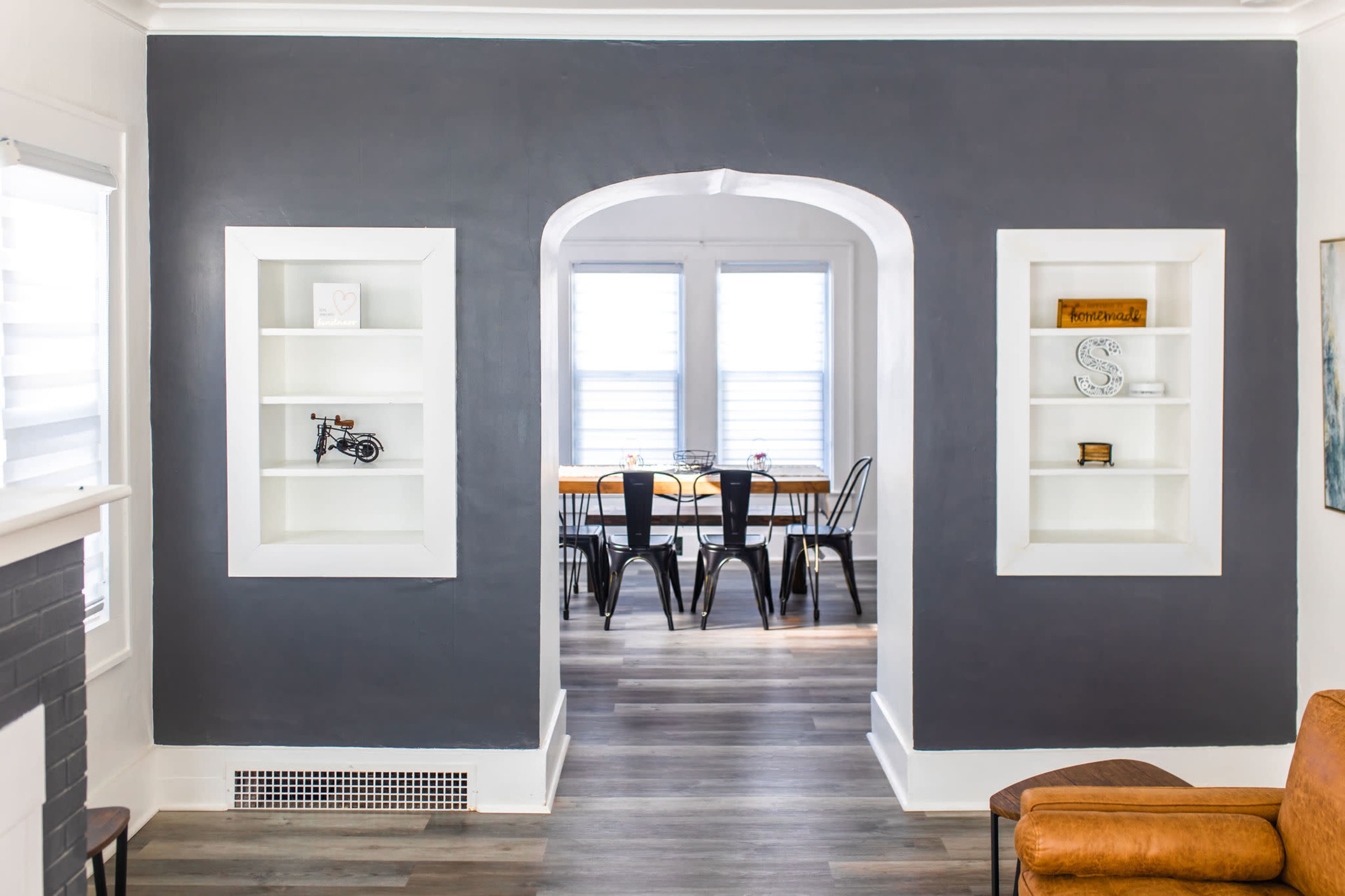 A living space with a dark gray wall featuring two white built-in shelves, leading to a dining area with a wooden table and black chairs.