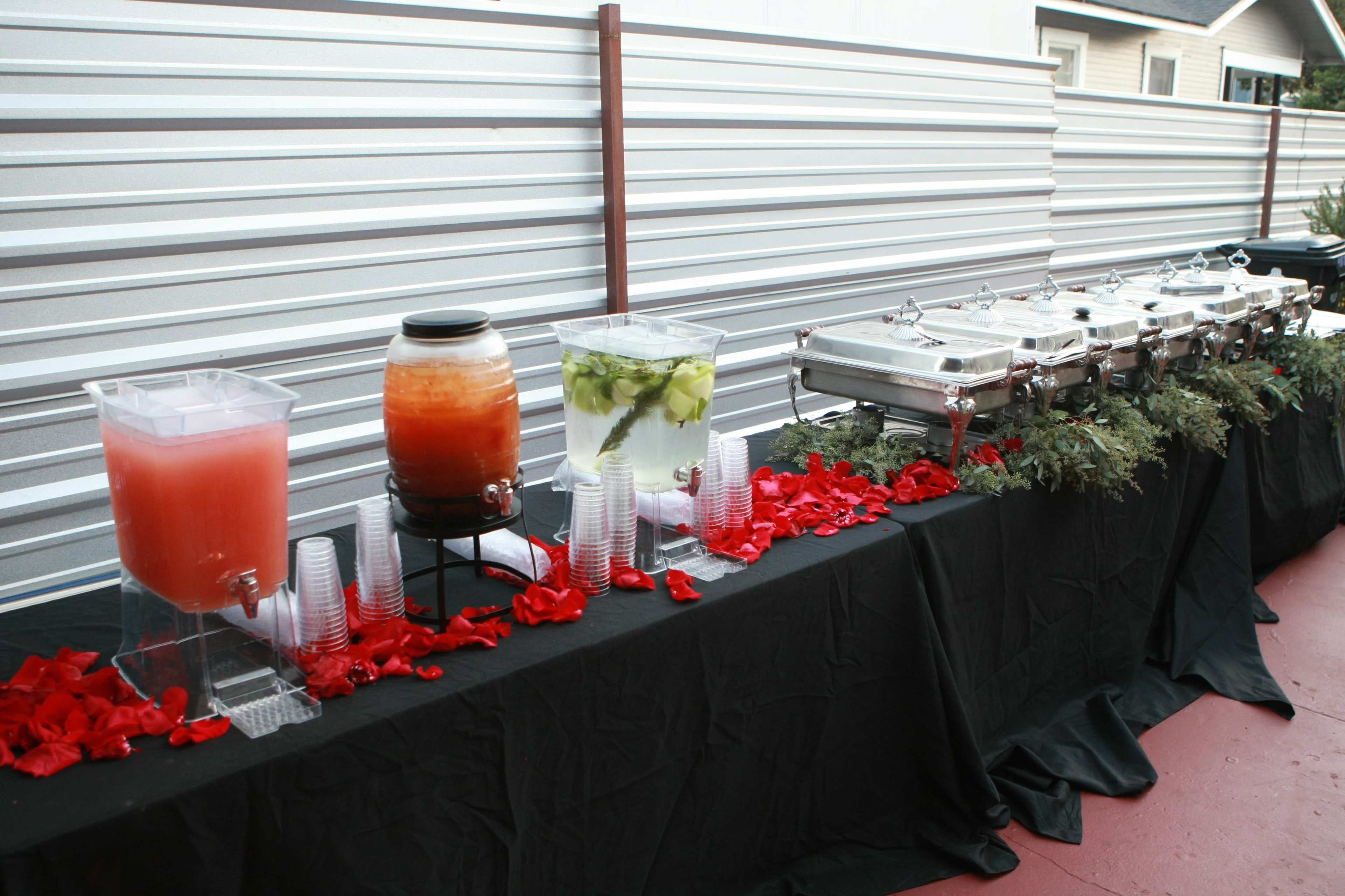 A long buffet table is set with two beverage dispensers and multiple food trays, adorned with red rose petals and greenery.