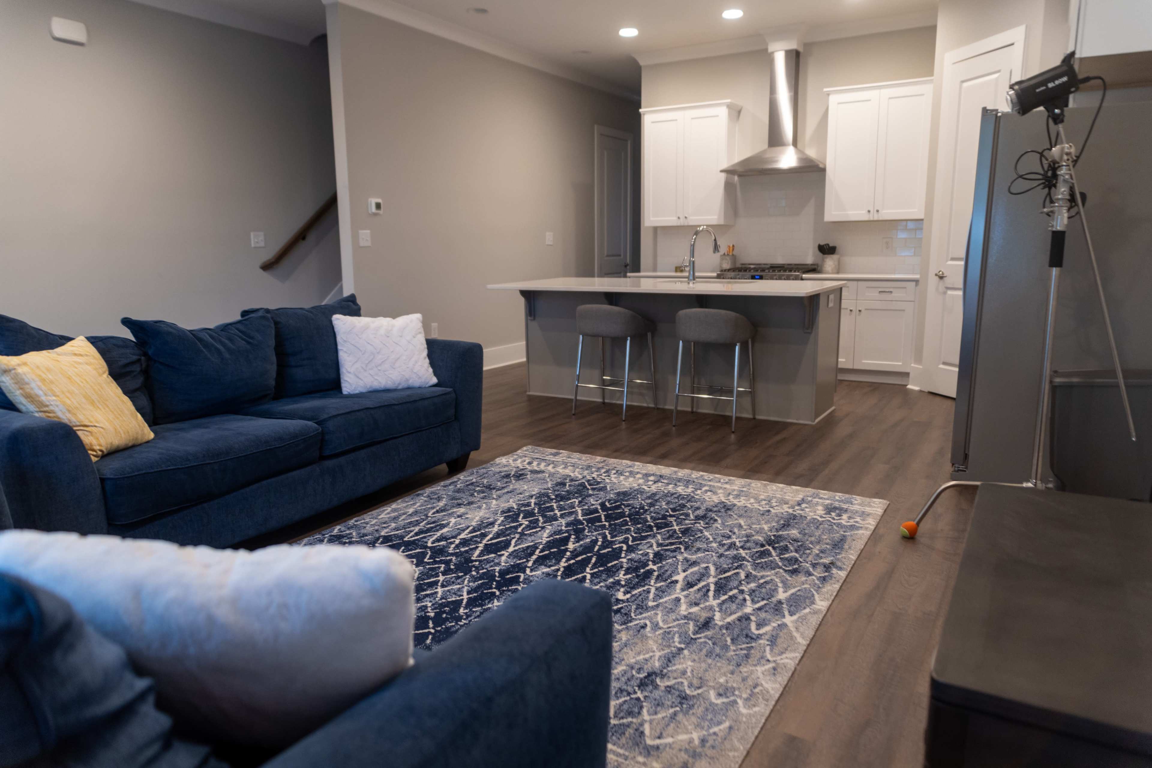 The image depicts a modern living room featuring a blue sectional sofa, a light blue rug, a kitchen island with two stools, and stainless steel appliances.