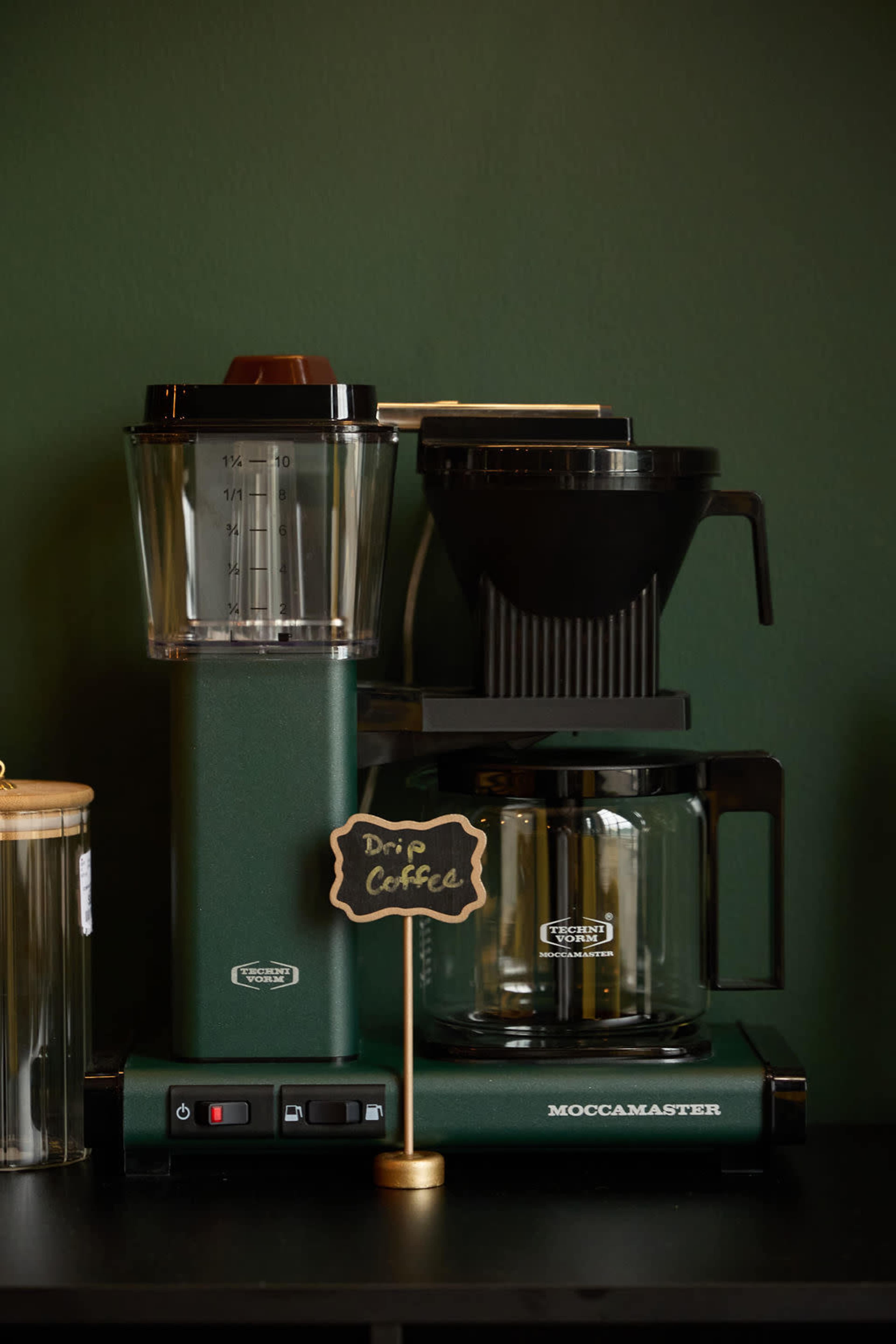 A green Moccamaster coffee maker with a glass carafe and a drip coffee filter sits on a dark countertop, accompanied by a small sign.
