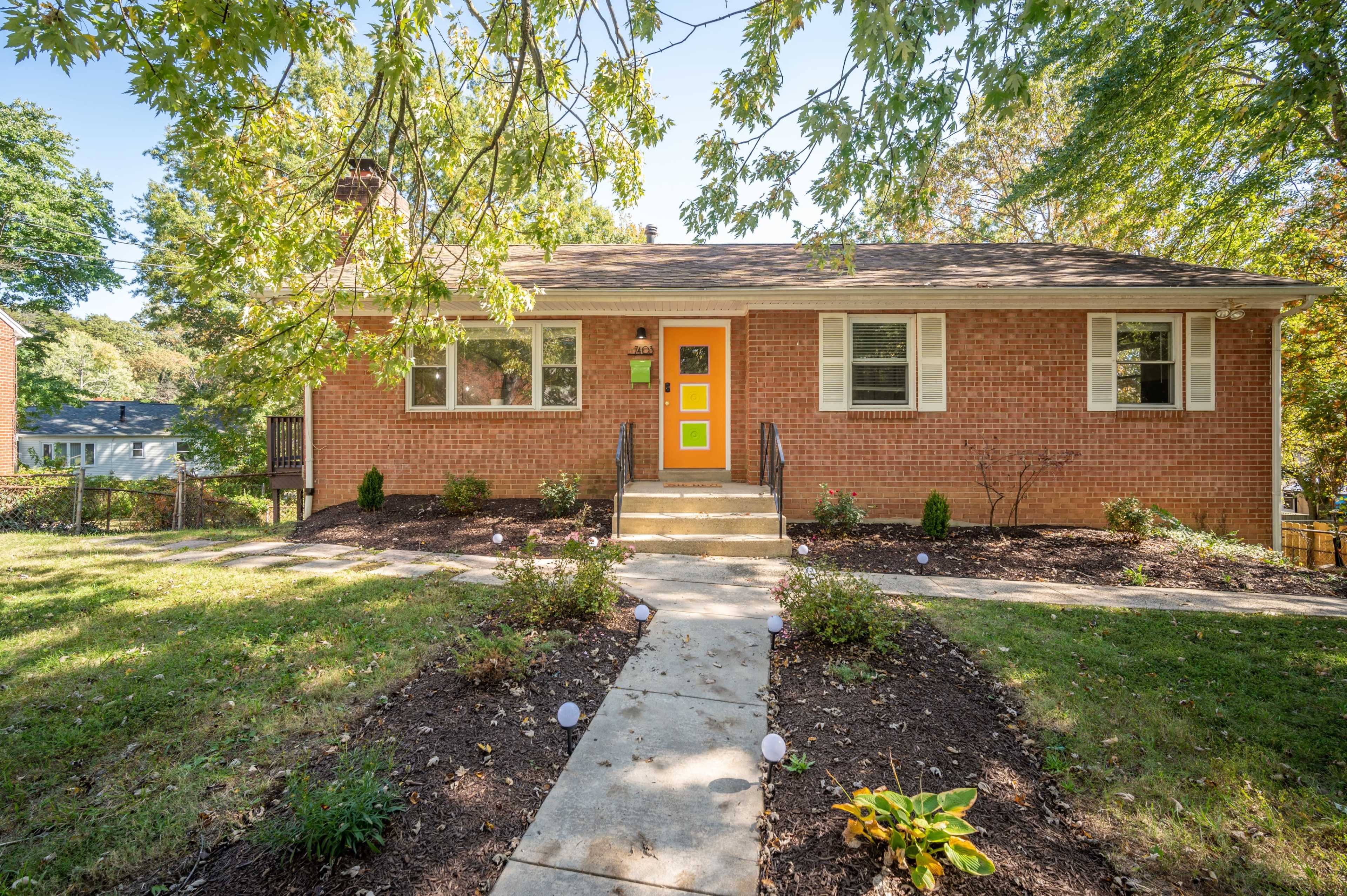 A brick house with an orange front door is flanked by green landscaping and a walkway leading to the entrance.