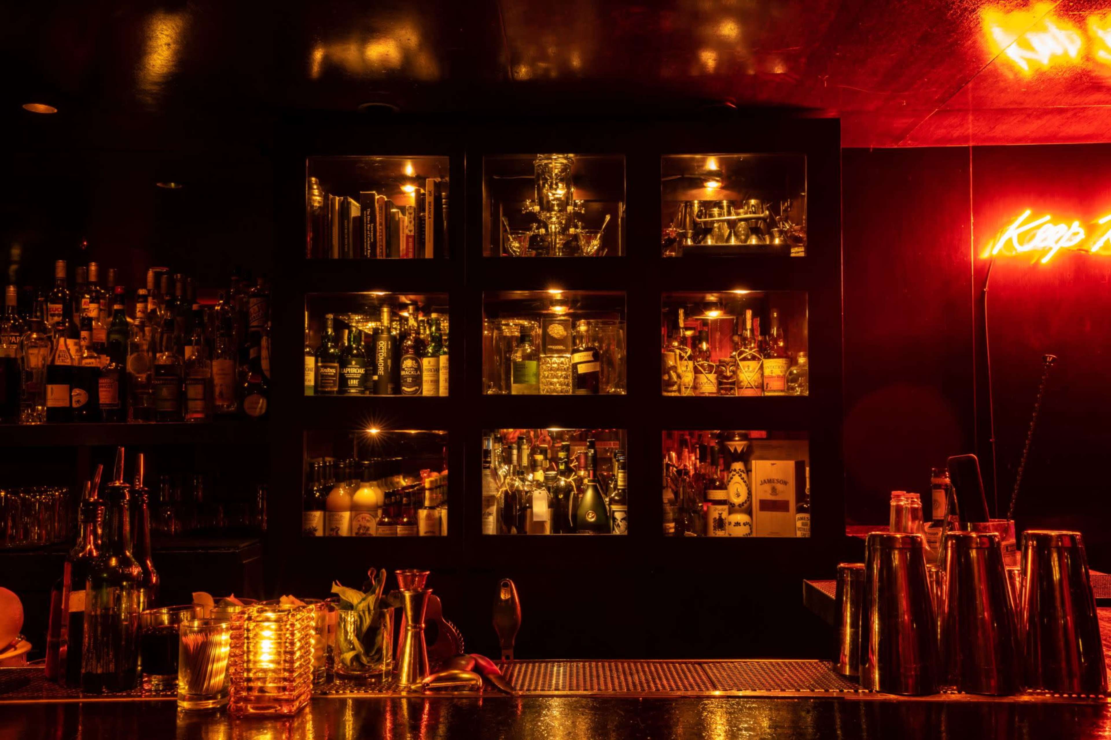 The image shows a dimly lit bar with shelves displaying various bottles of liquor illuminated by soft lighting, and a neon sign that reads "Keep."