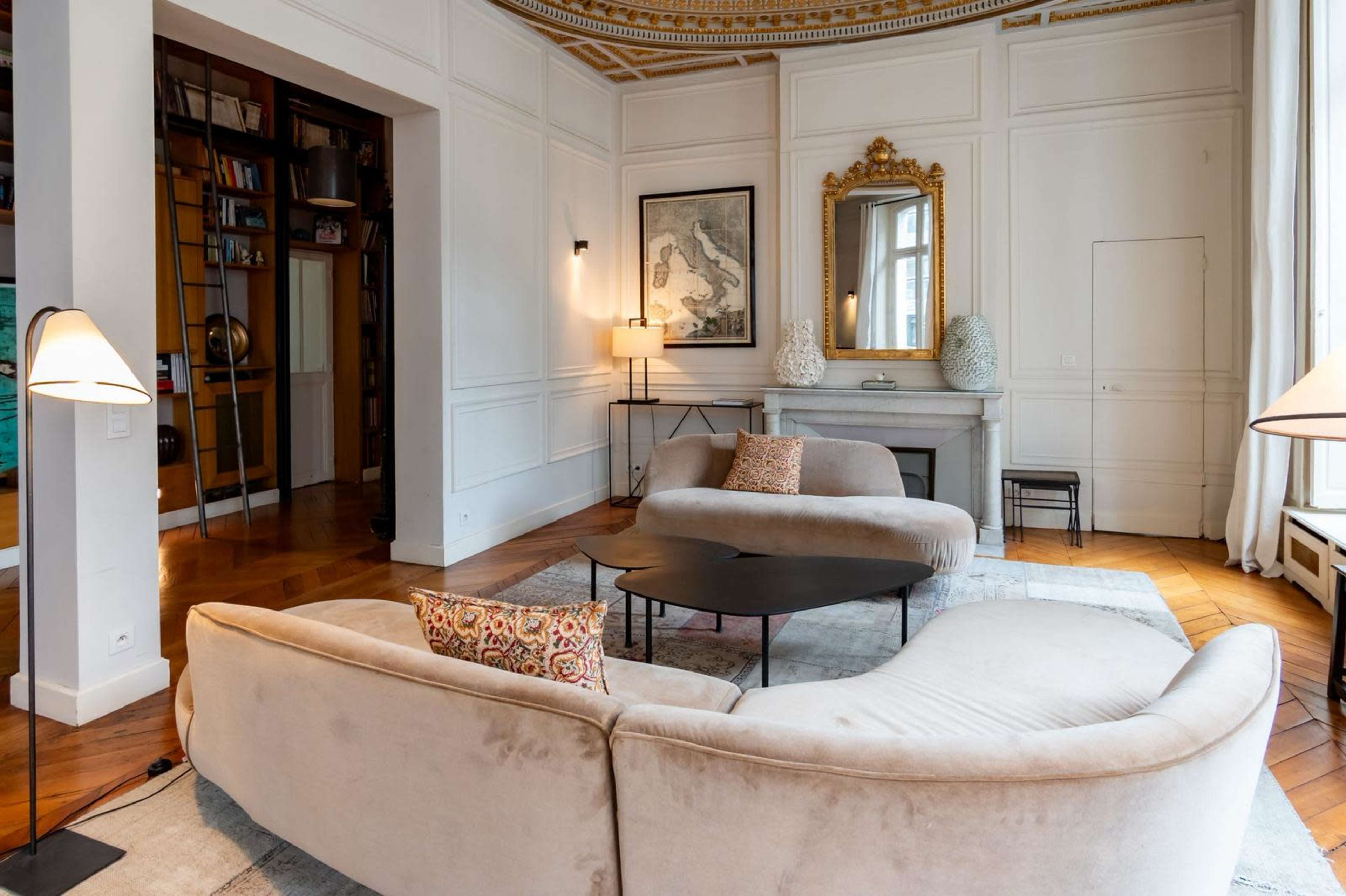 The image shows a spacious living room featuring a curved beige sofa, a black coffee table, and a fireplace with a decorative mirror above it, all set against an elegant backdrop of white walls and hardwood flooring.