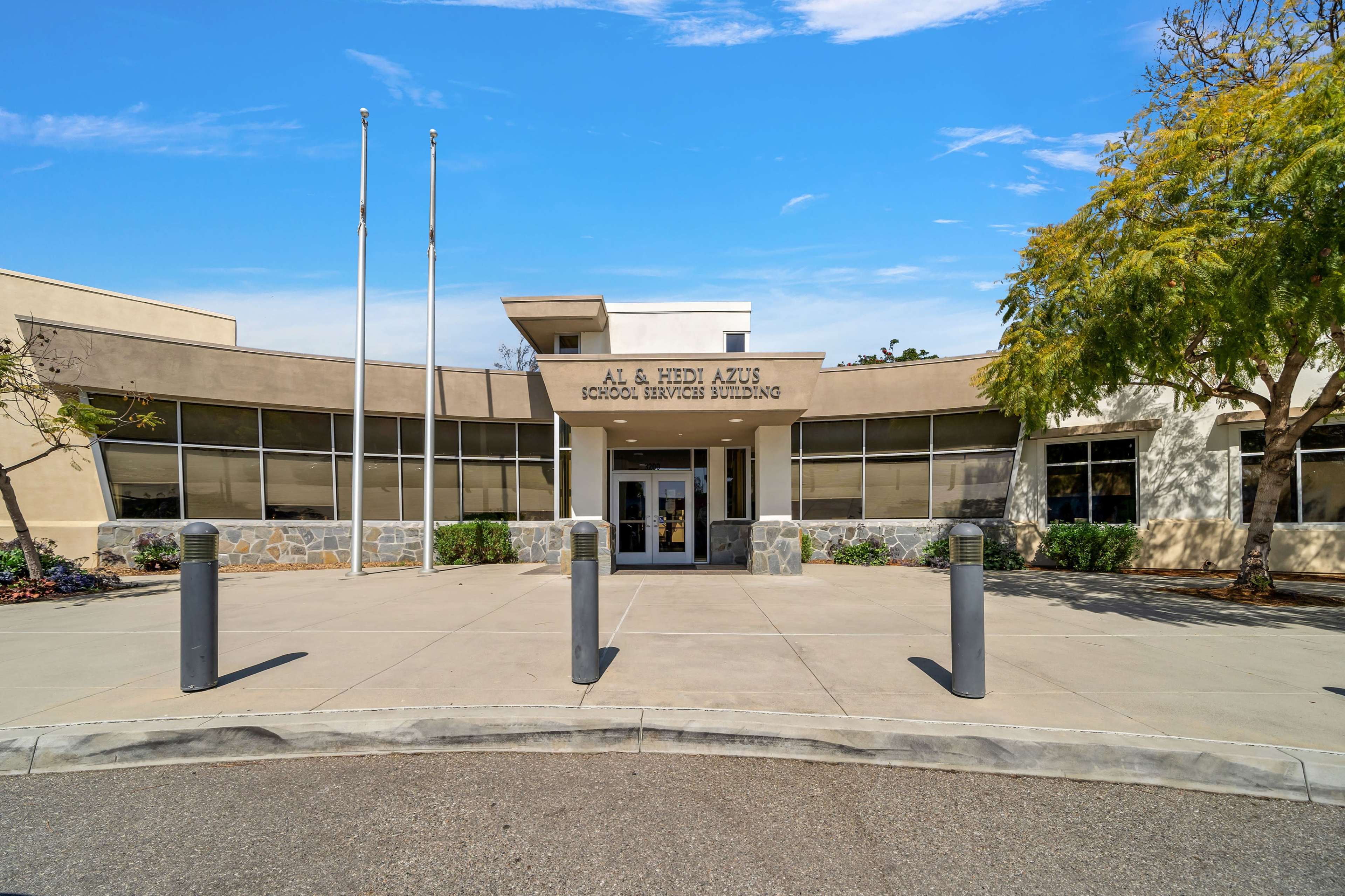 A modern school administration building features large front windows and a central entrance, flanked by flag poles.
