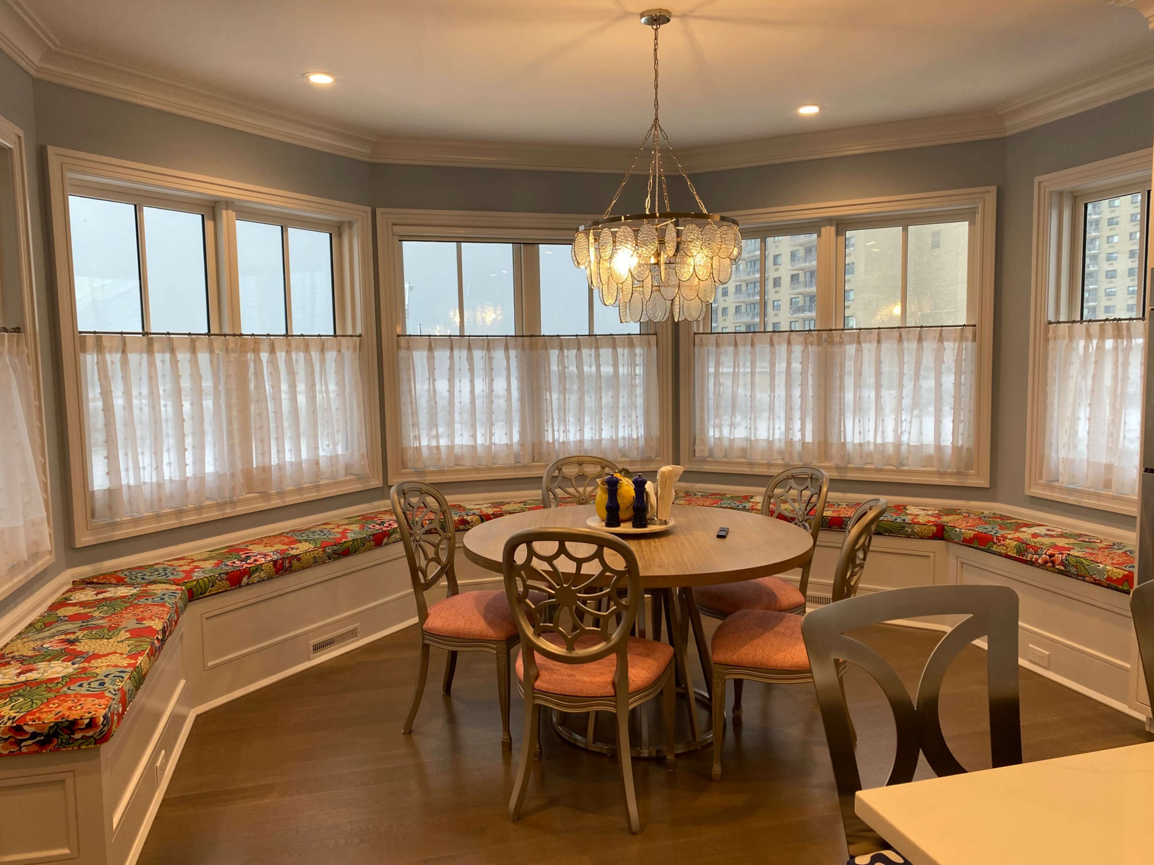 A dining area features a circular table surrounded by chairs, with colorful cushions lining a bay window illuminated by natural light.