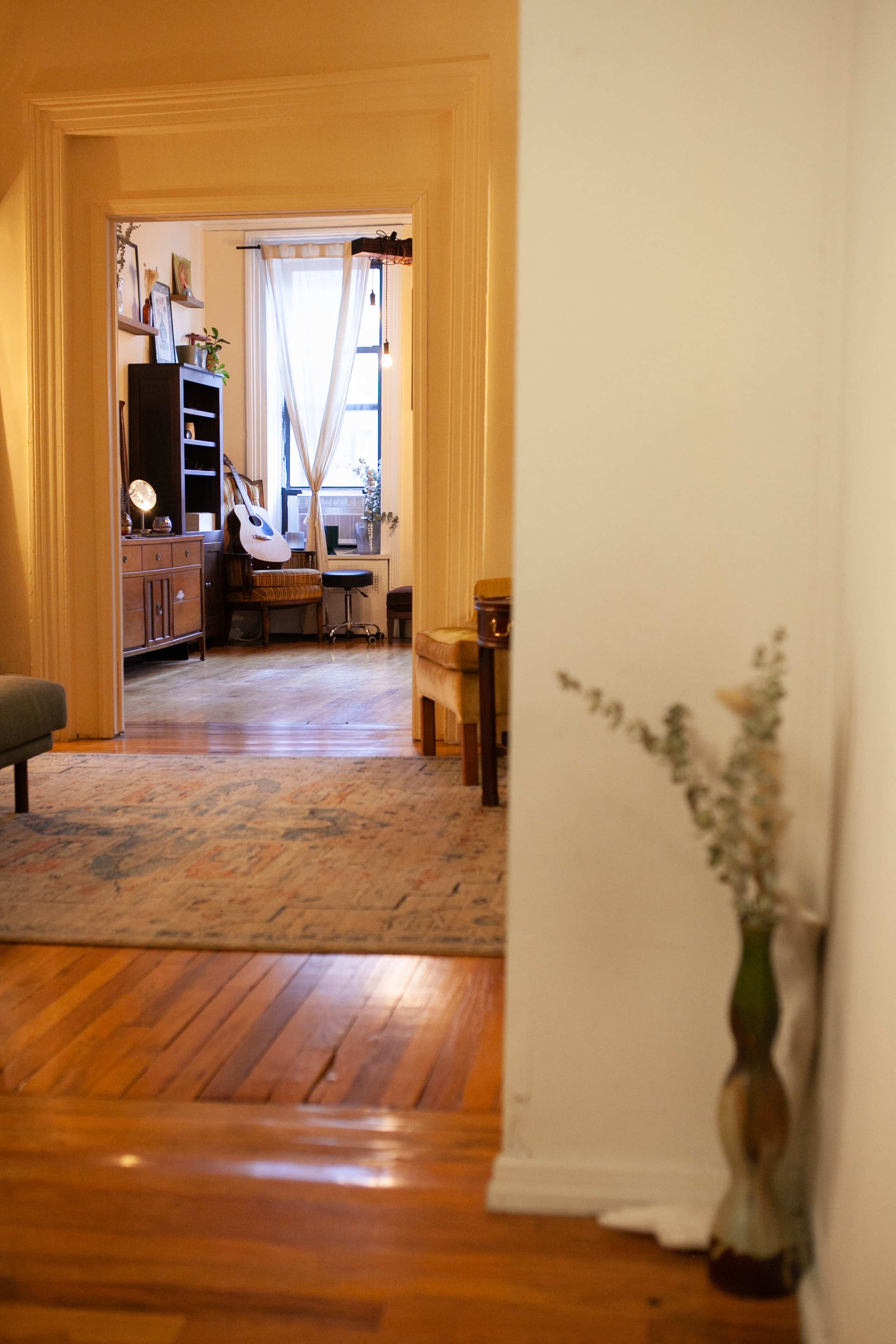 A hallway leads to a cozy living room with wooden floors, a large window, and various furniture pieces.