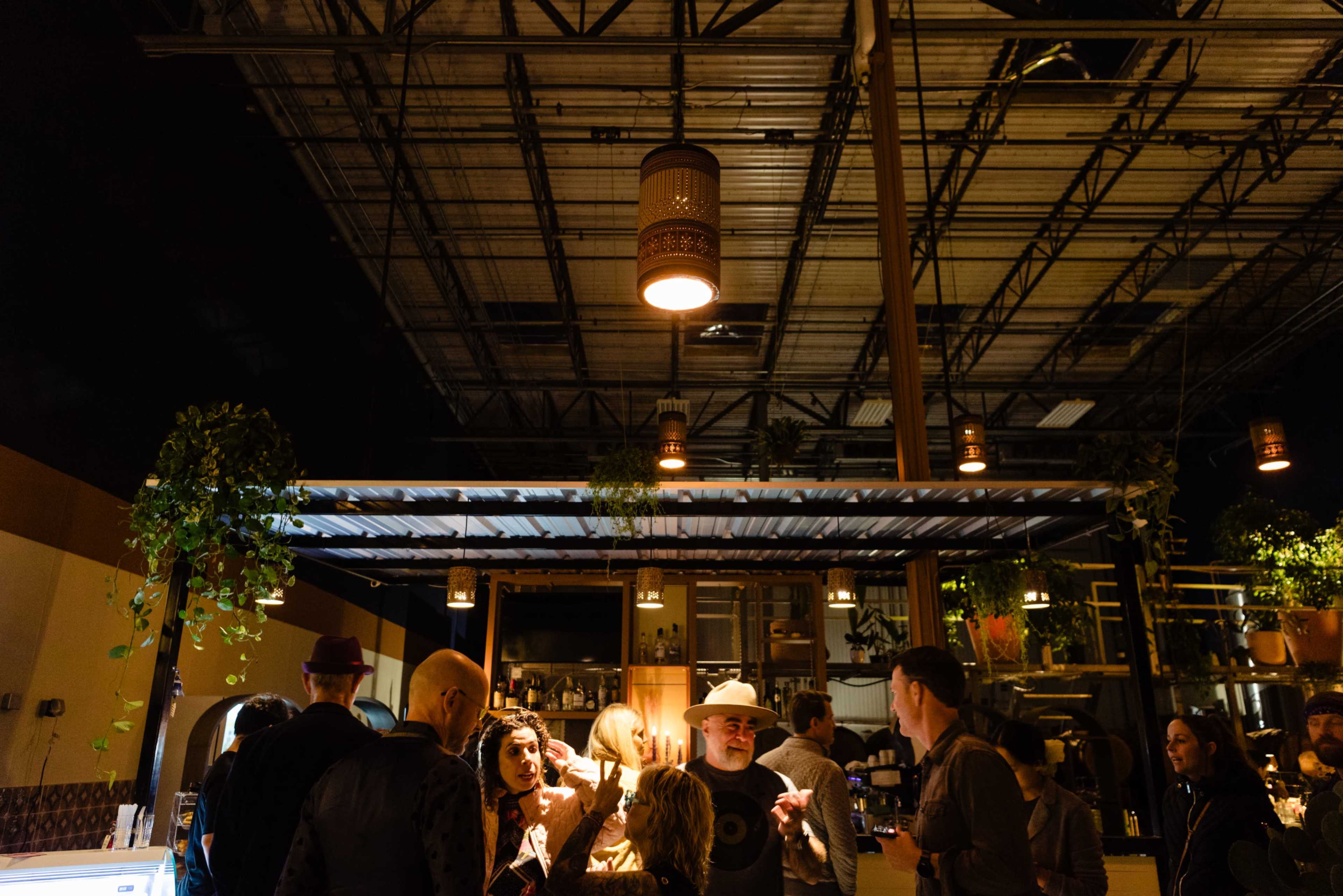 A group of people socializes at a bar in an industrial-style venue with overhead lights and greenery.