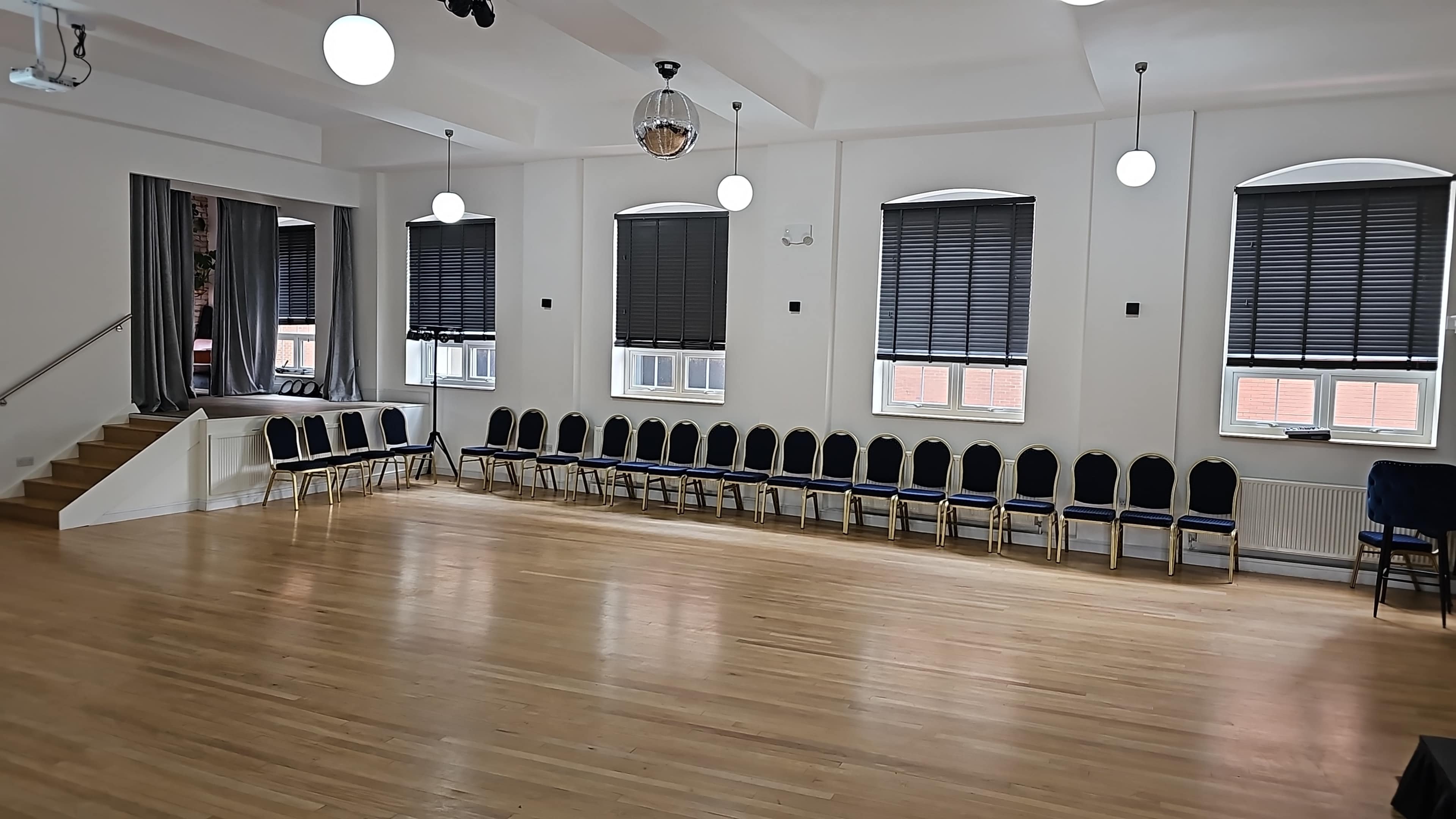 The image shows a spacious room with a polished wooden floor, featuring rows of blue chairs along one wall and large windows covered with dark blinds.