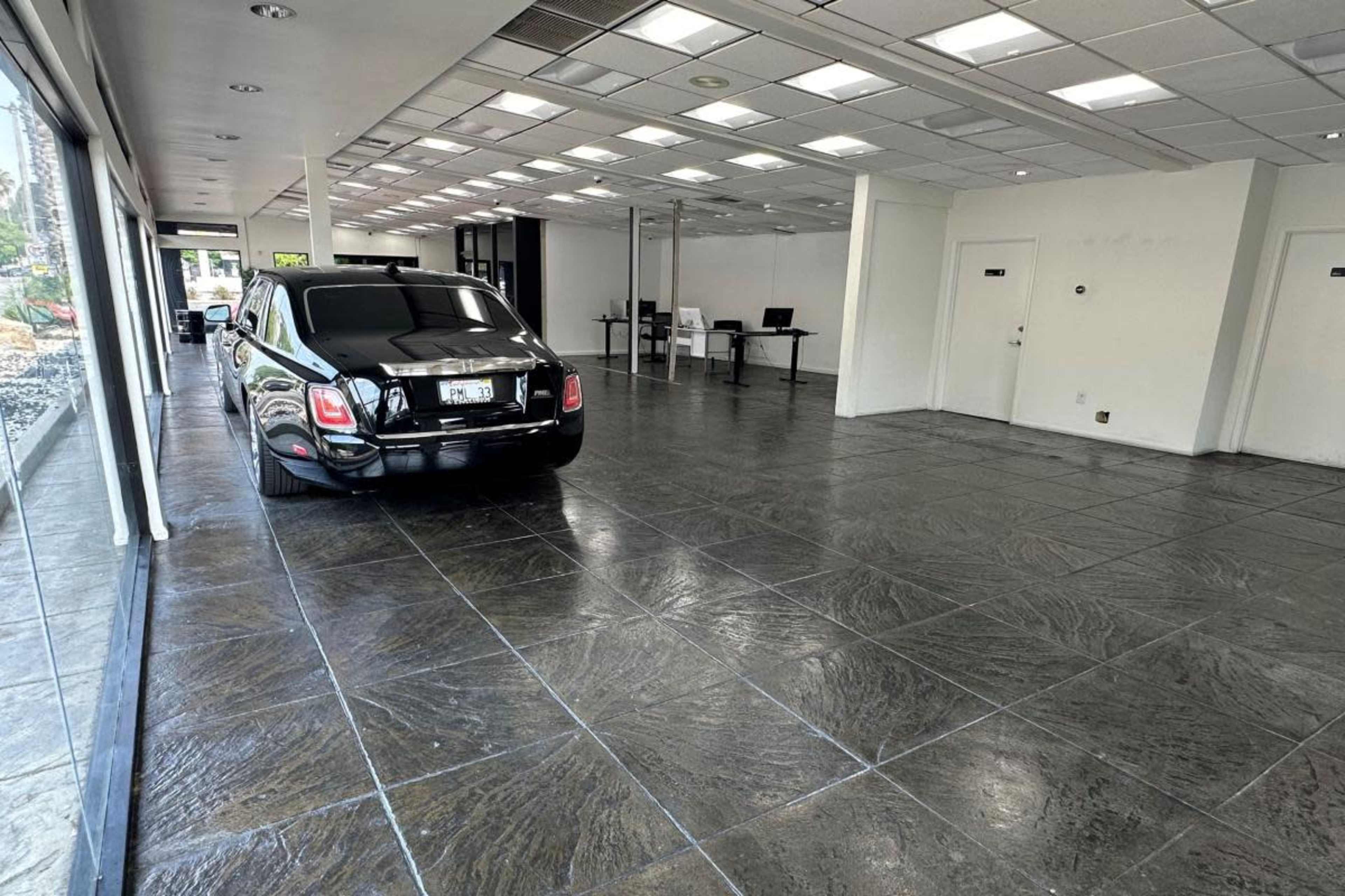 The image shows a spacious, empty room with a polished stone floor, featuring a black car parked on one side and several tables set up for work on the opposite side.
