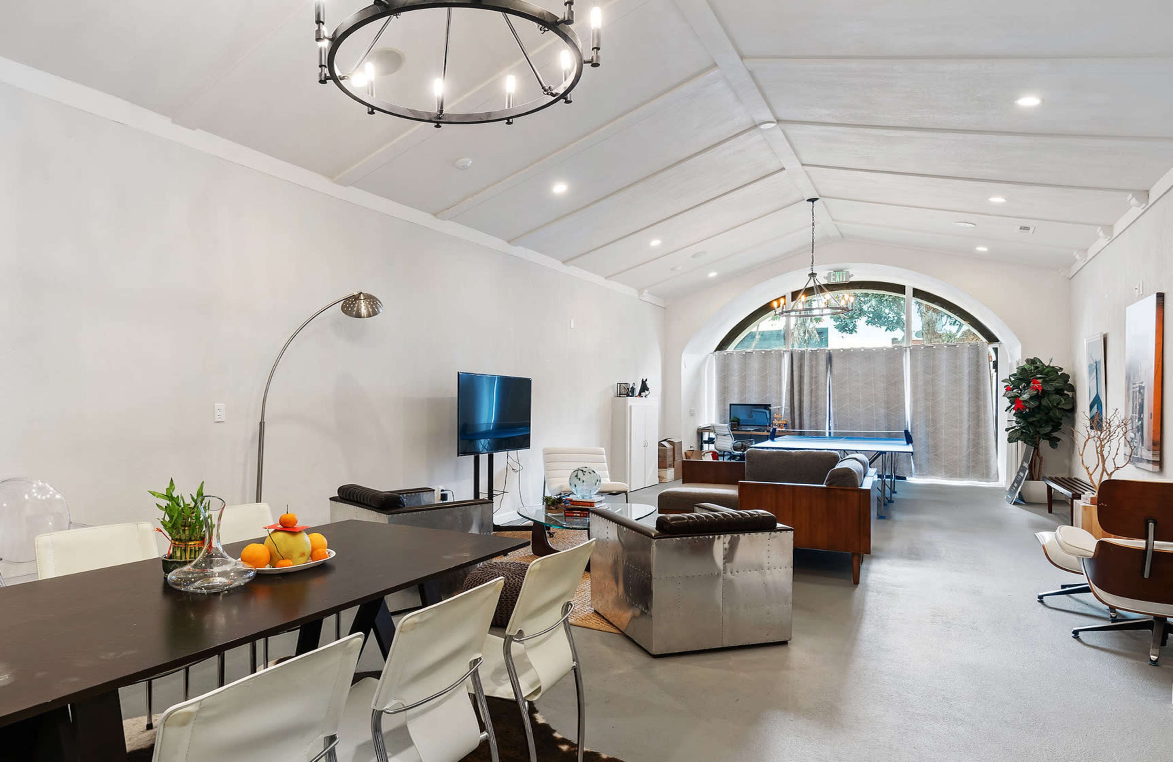 A spacious living area with a dining table, a television, and a seating arrangement, featuring high ceilings and large windows.