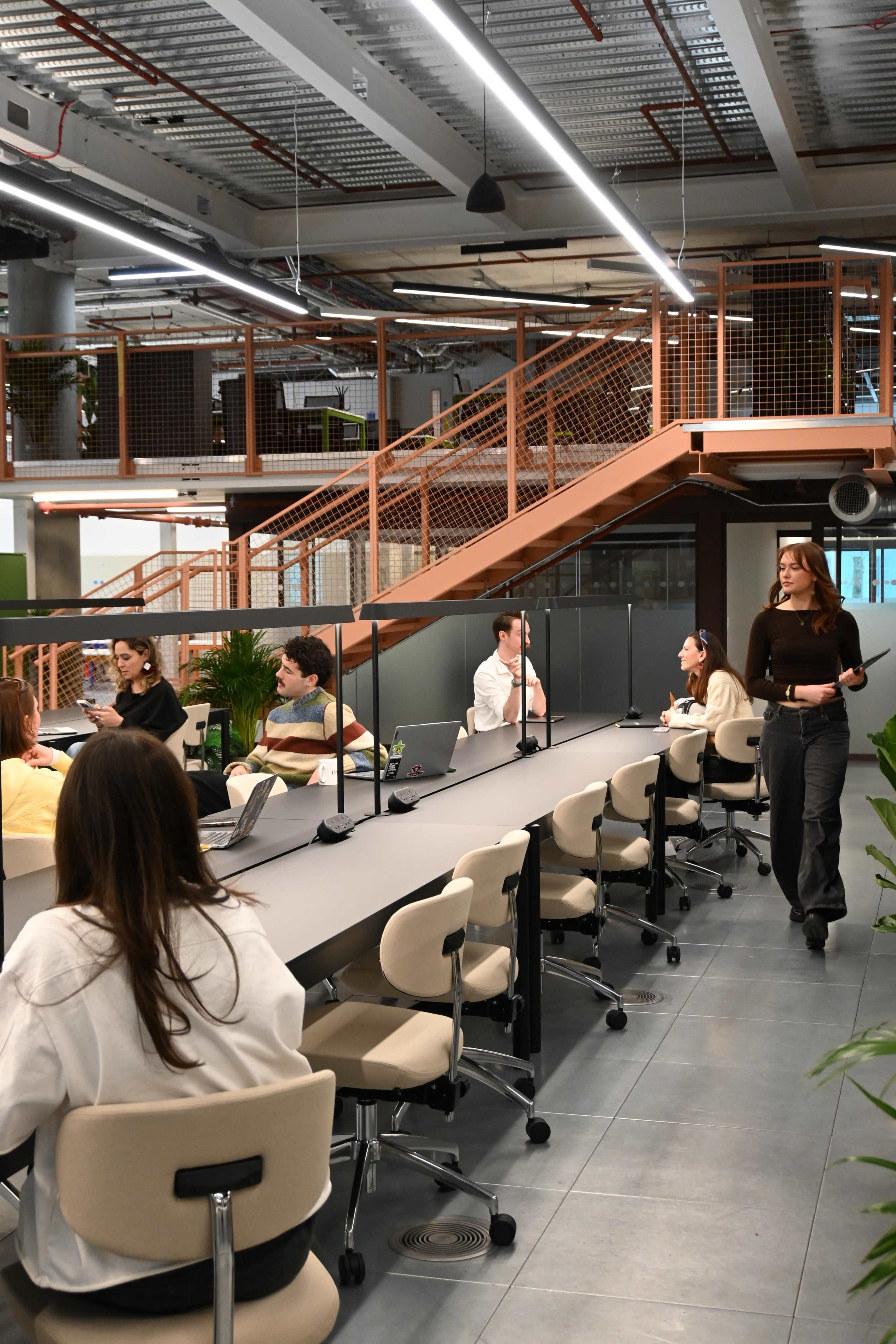 The image shows a modern office space with multiple workstations, where several people are engaged in discussions while others work on laptops.