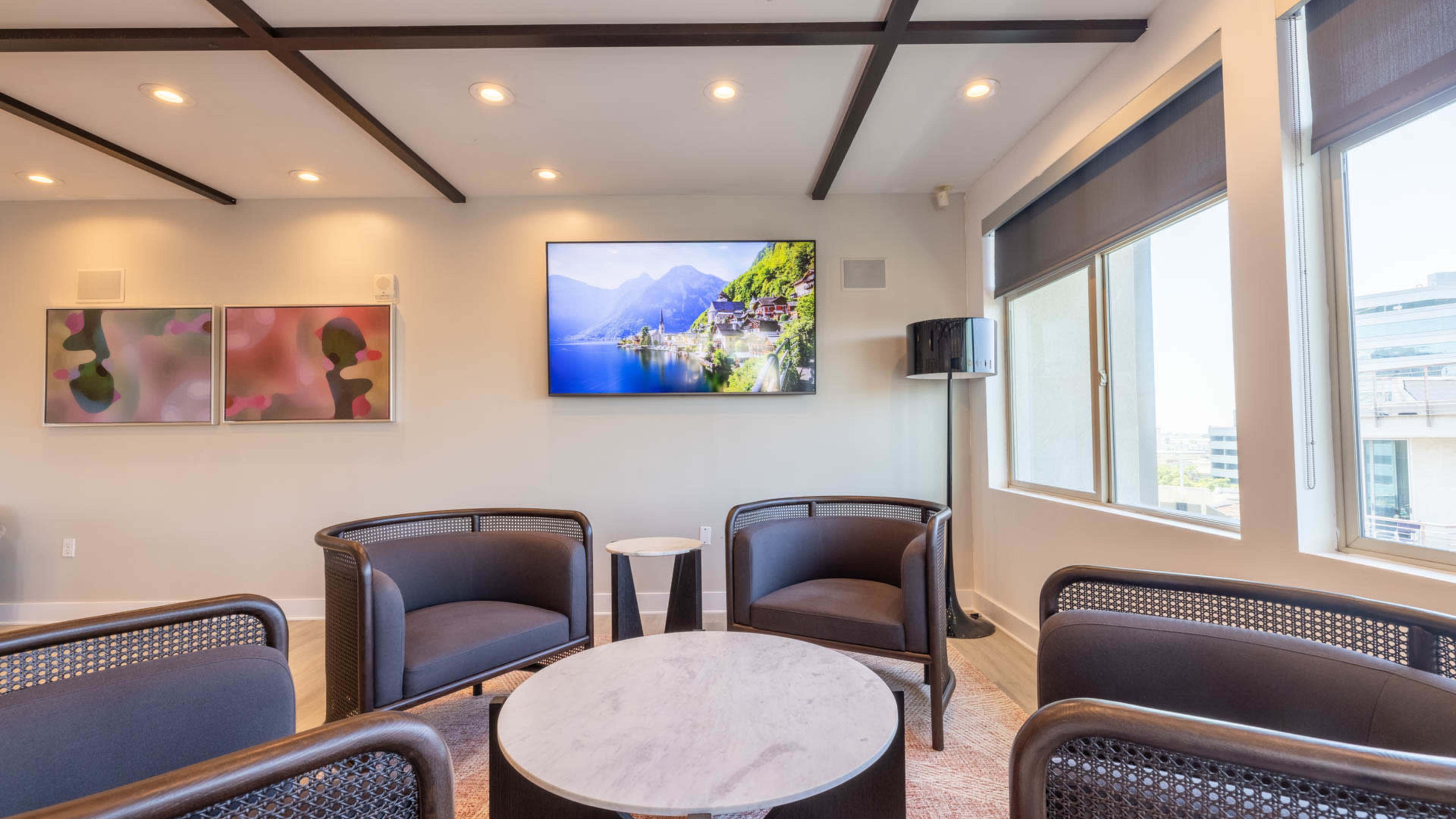 The image shows a modern, well-lit lounge area with four dark wicker chairs arranged around a round marble table, a large TV display mounted on the wall, and scenic artwork and windows allowing natural light.