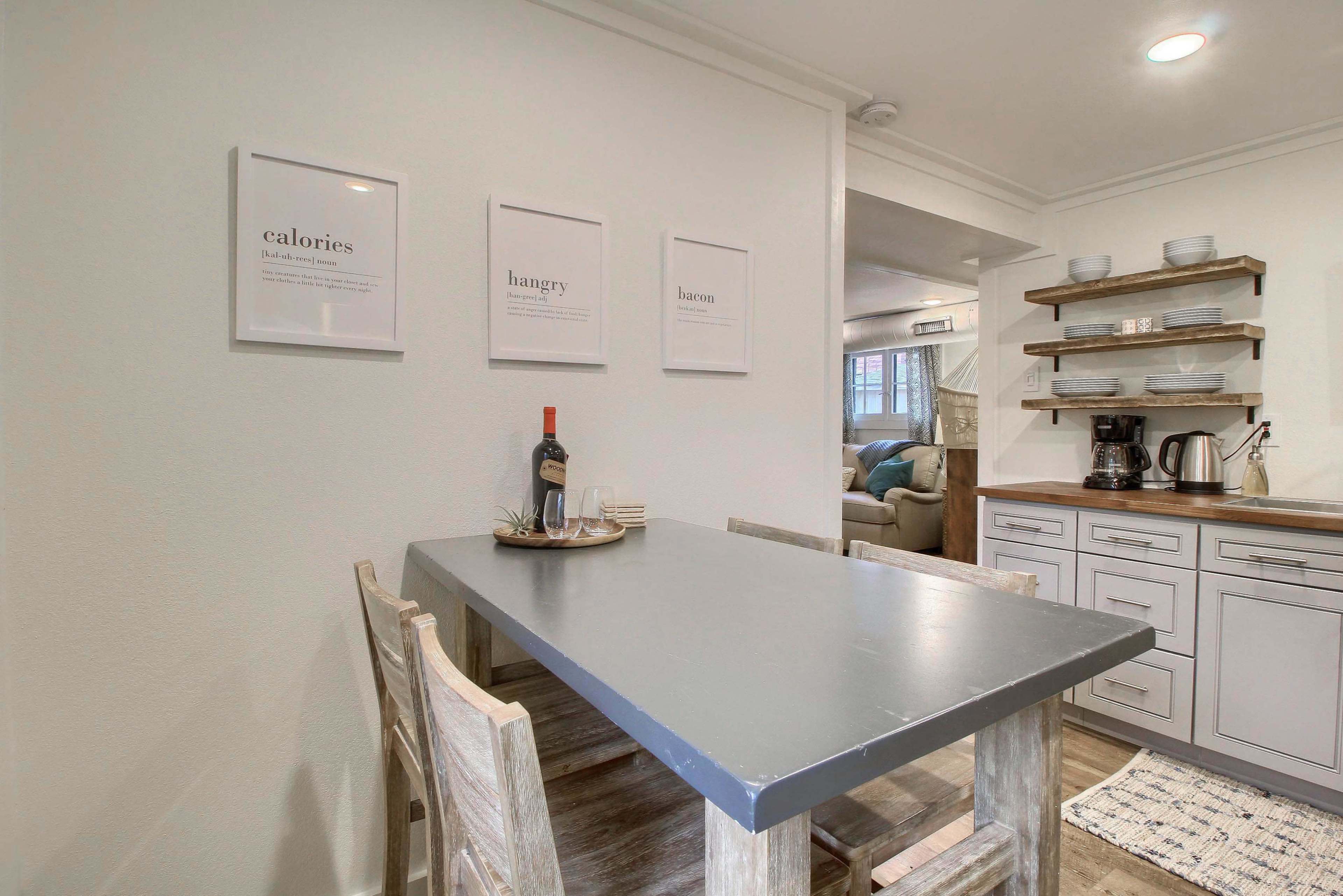A simple kitchen area features a gray table with wooden chairs, framed prints on the wall, and neatly arranged kitchenware on a shelf.