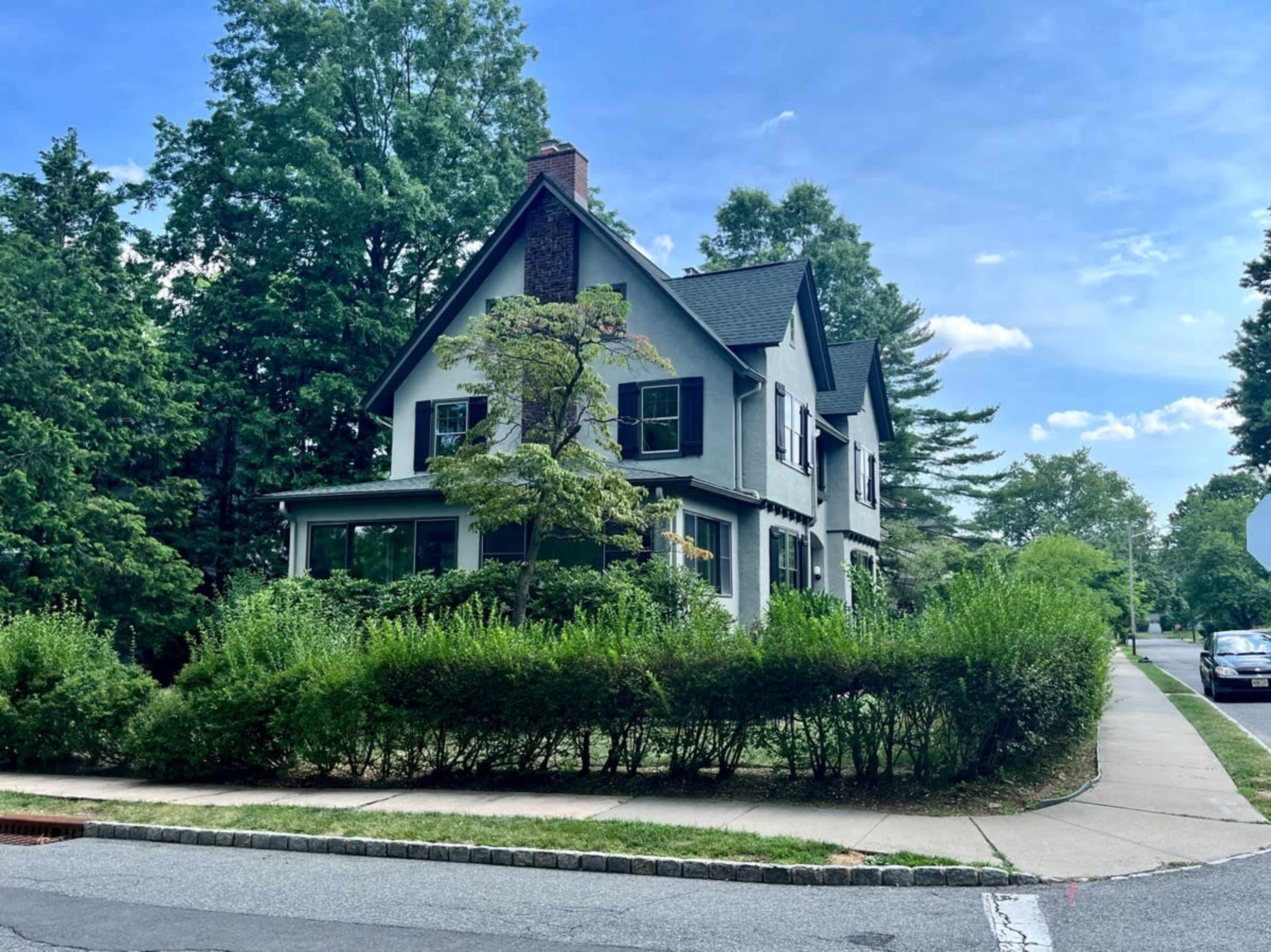 A two-story house with a gabled roof is surrounded by green shrubs and trees on a residential street.