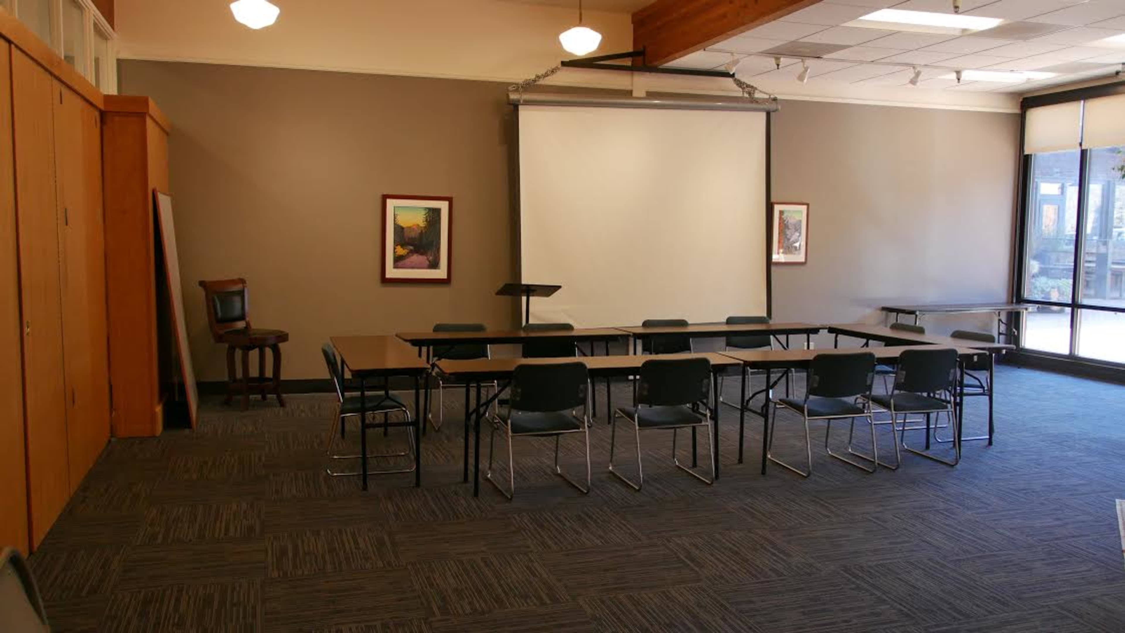 A meeting room features a central arrangement of tables and chairs, with a projector screen and framed artwork on the walls.