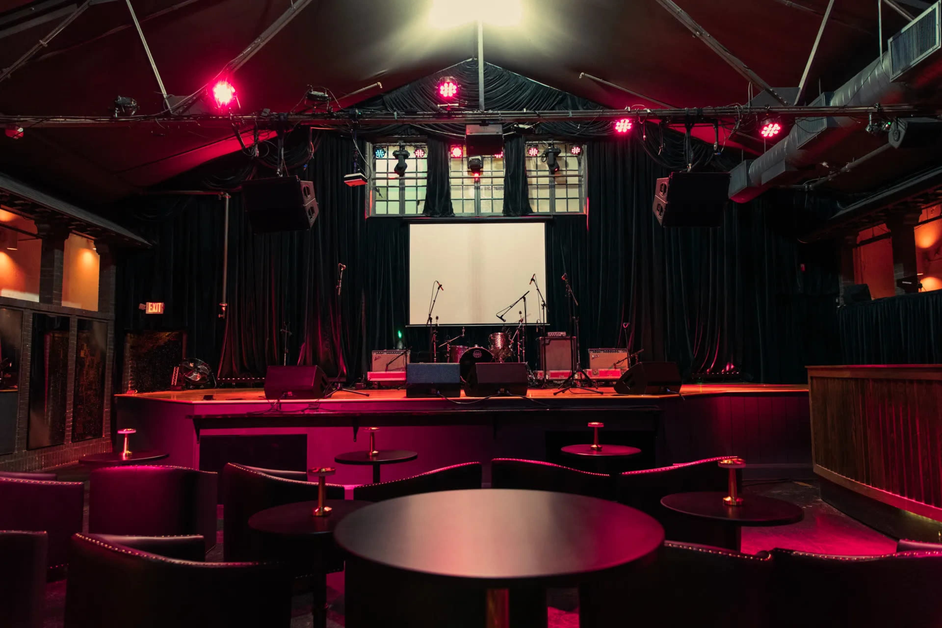 The image shows an empty stage in a dimly lit venue with musical instruments set up and tables in the foreground.