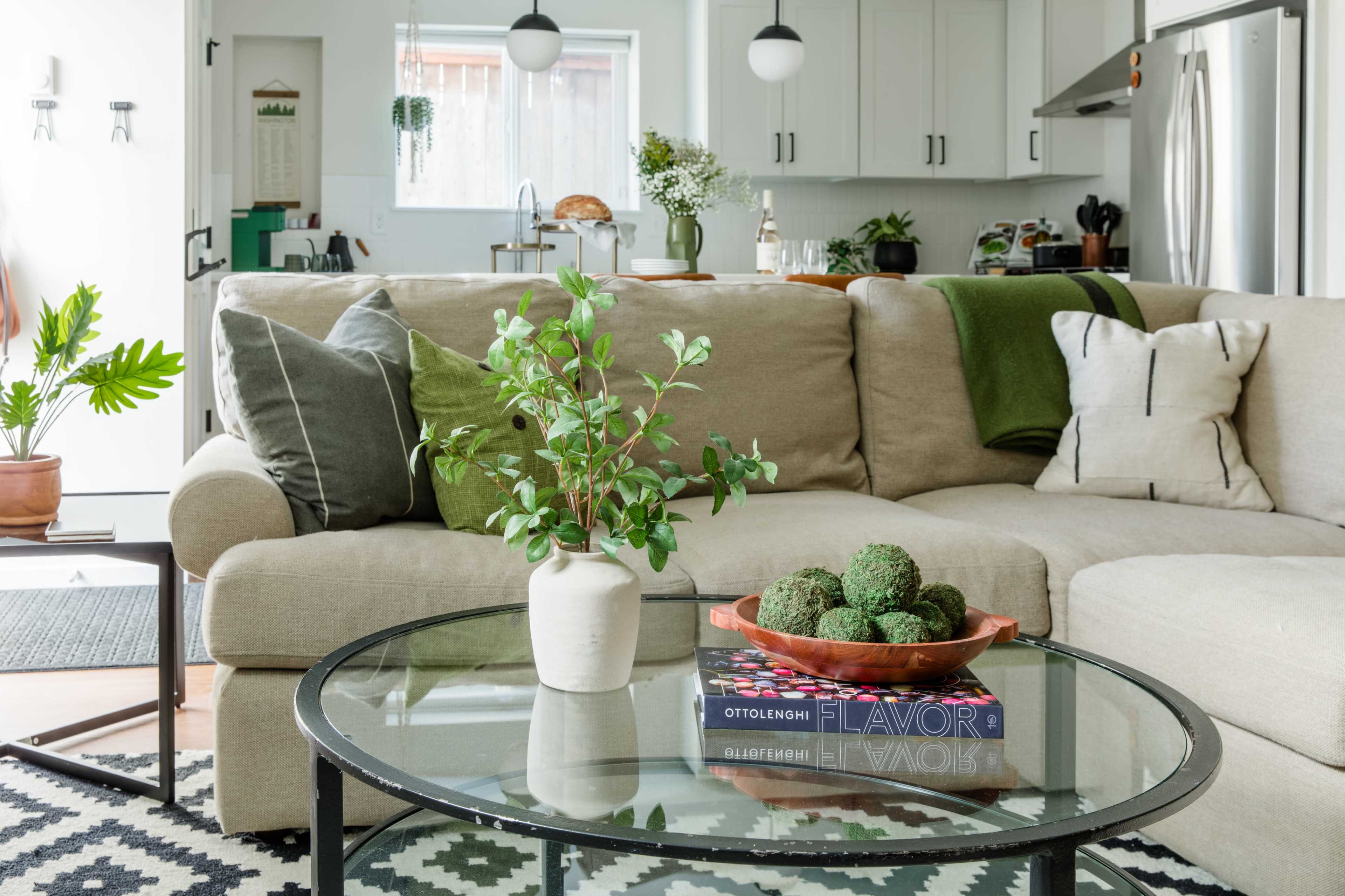 A cozy living room features a light-colored sectional sofa adorned with green throw pillows, a glass coffee table with decorative items, and a plant in a vase.