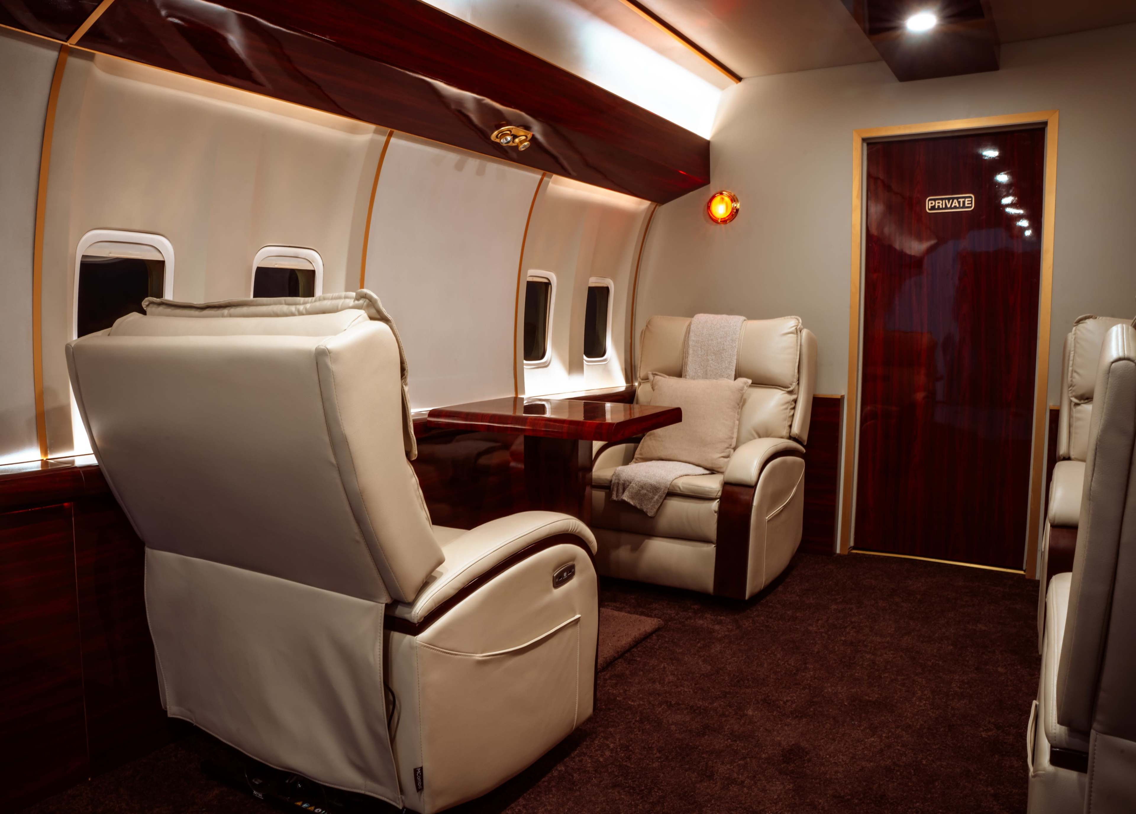 The image shows the interior of a private jet featuring two plush armchairs and a table, with a door labeled "PRIVATE" at the back.